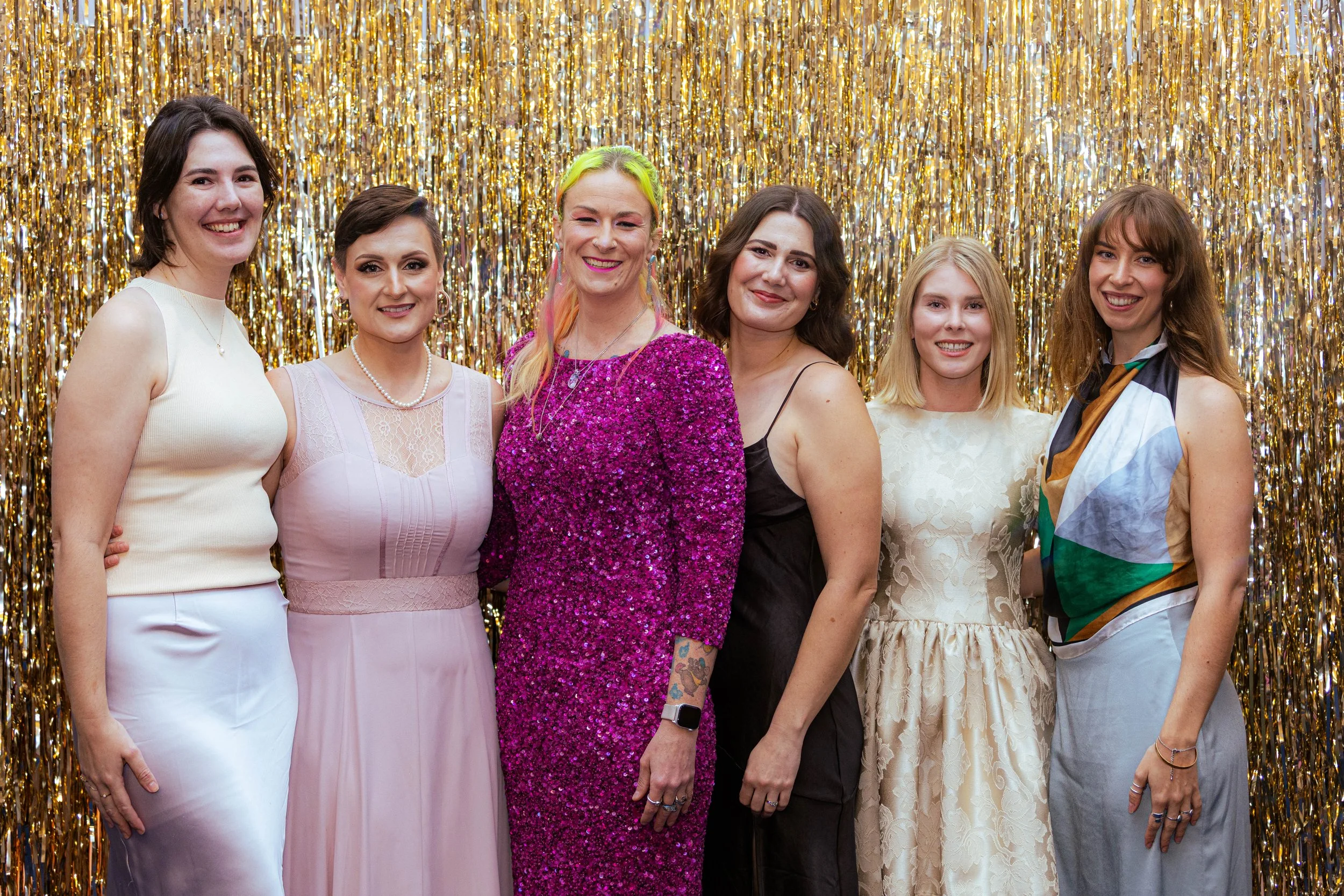 A group of seven women standing in front of a gold tinsel backdrop, dressed in stylish outfits, smiling and posing for a photo.