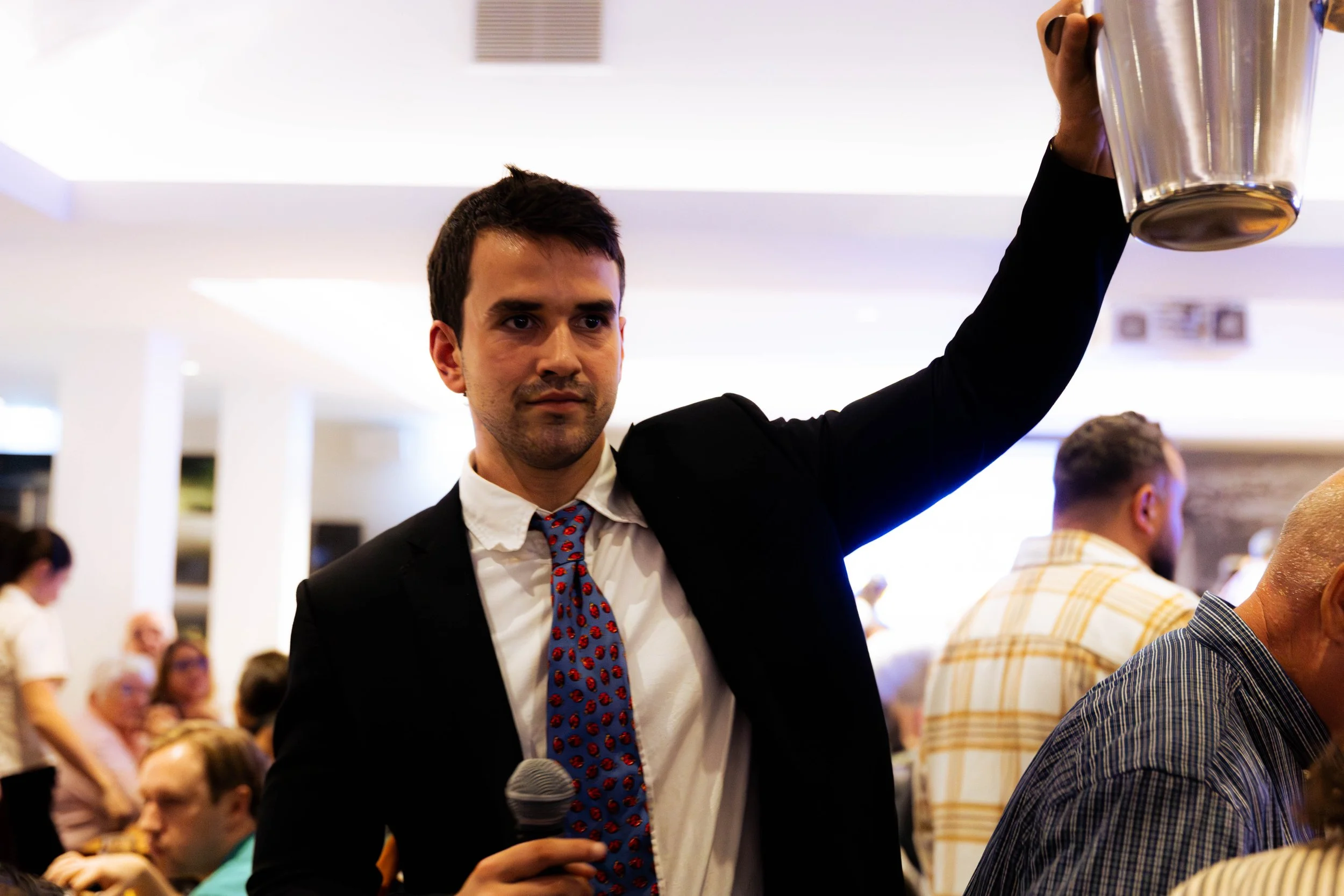 A man in a black suit, white shirt, and colorful tie holding a microphone in a large indoor gathering. He is raising a glass in a toast with a serious expression. The background features a group of people seated and standing, some focused on the man.