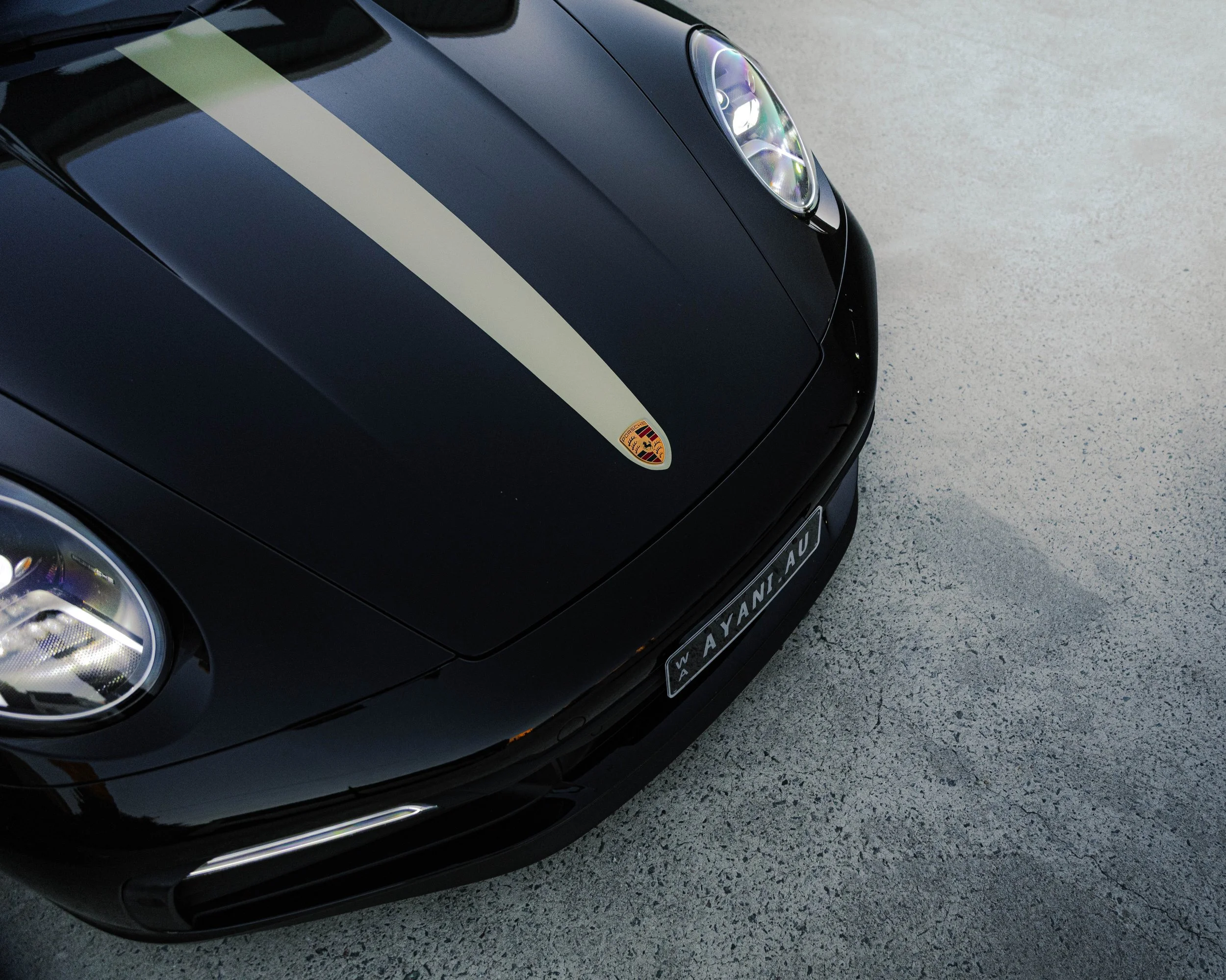 Close-up of a black sports car with a Porsche emblem, a white racing stripe on the hood, and a European-style license plate, parked on a light gray concrete surface.