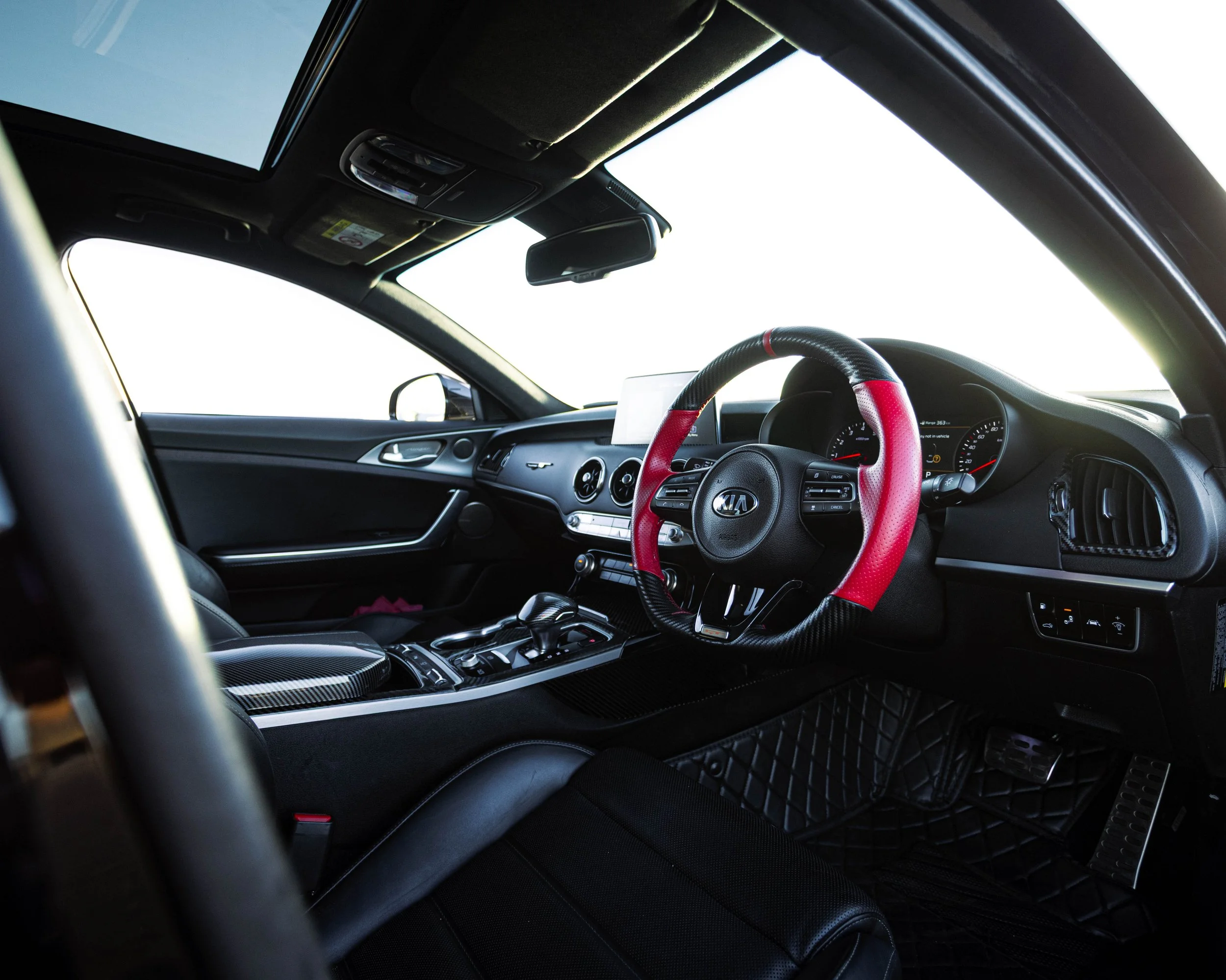 Interior of a car showing the steering wheel with a red and black cover, dashboard, rearview mirror, and sunroof from the passenger side view.