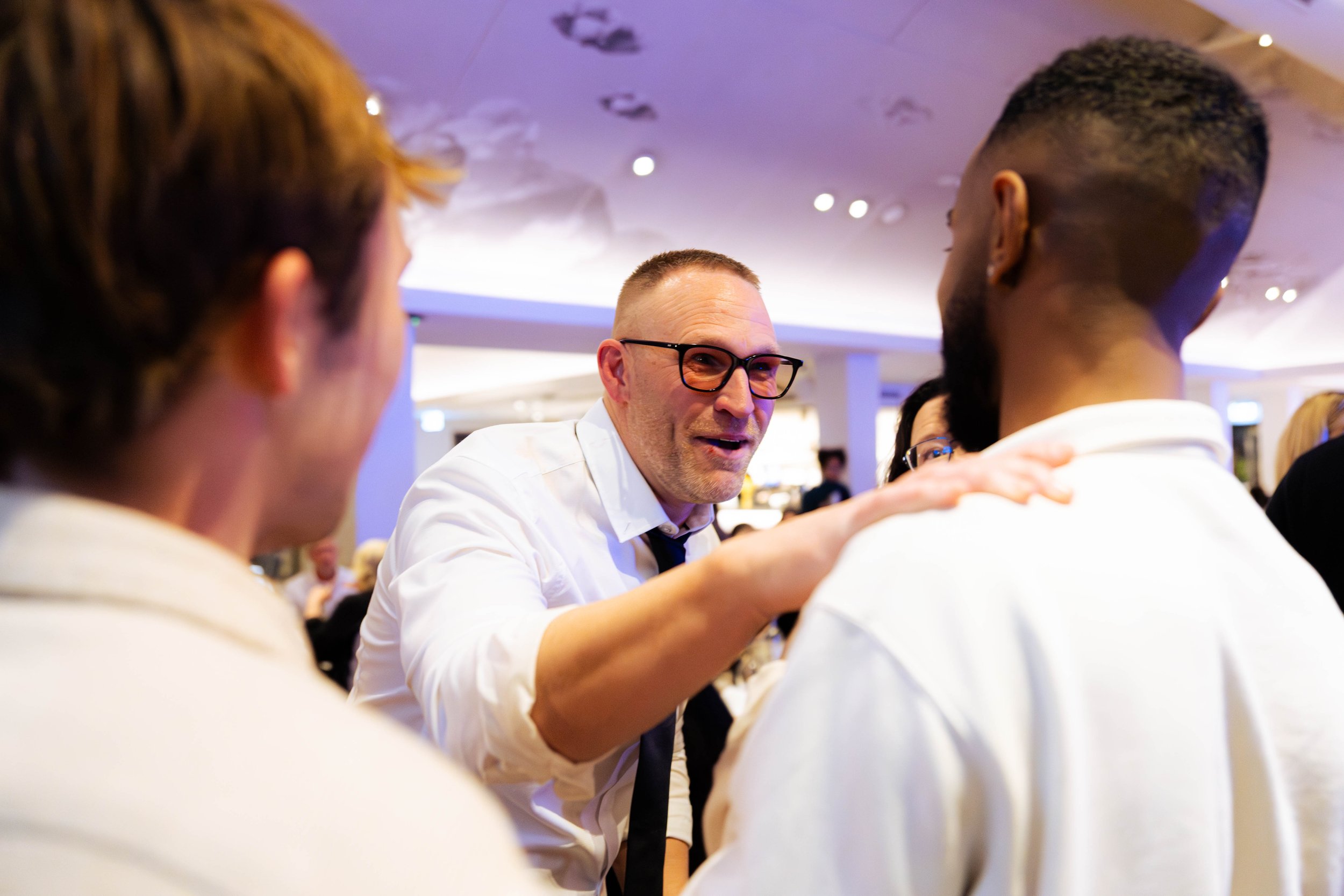 Three men in conversation at a social gathering, with one man in the center wearing glasses and a white shirt smiling while others listen.