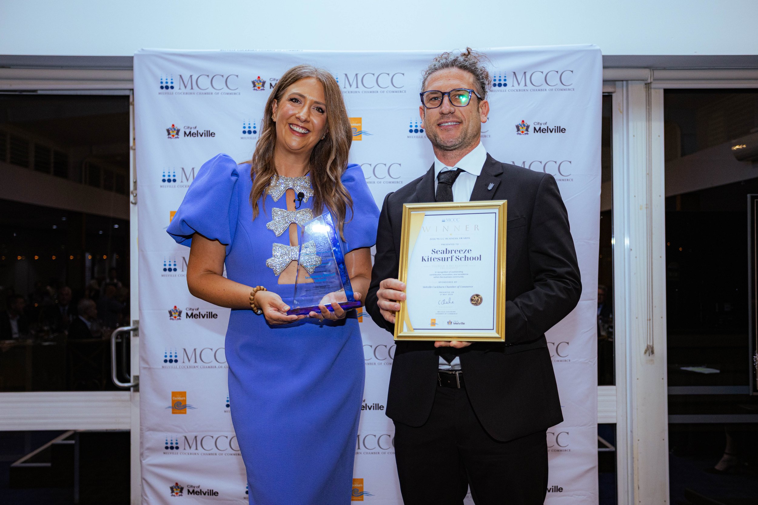 A woman in a blue dress and a man in a black suit and glasses are standing side by side, smiling at an event. The woman is holding a glass award, and the man is holding a framed certificate. The background features a banner with logos and text, indic
