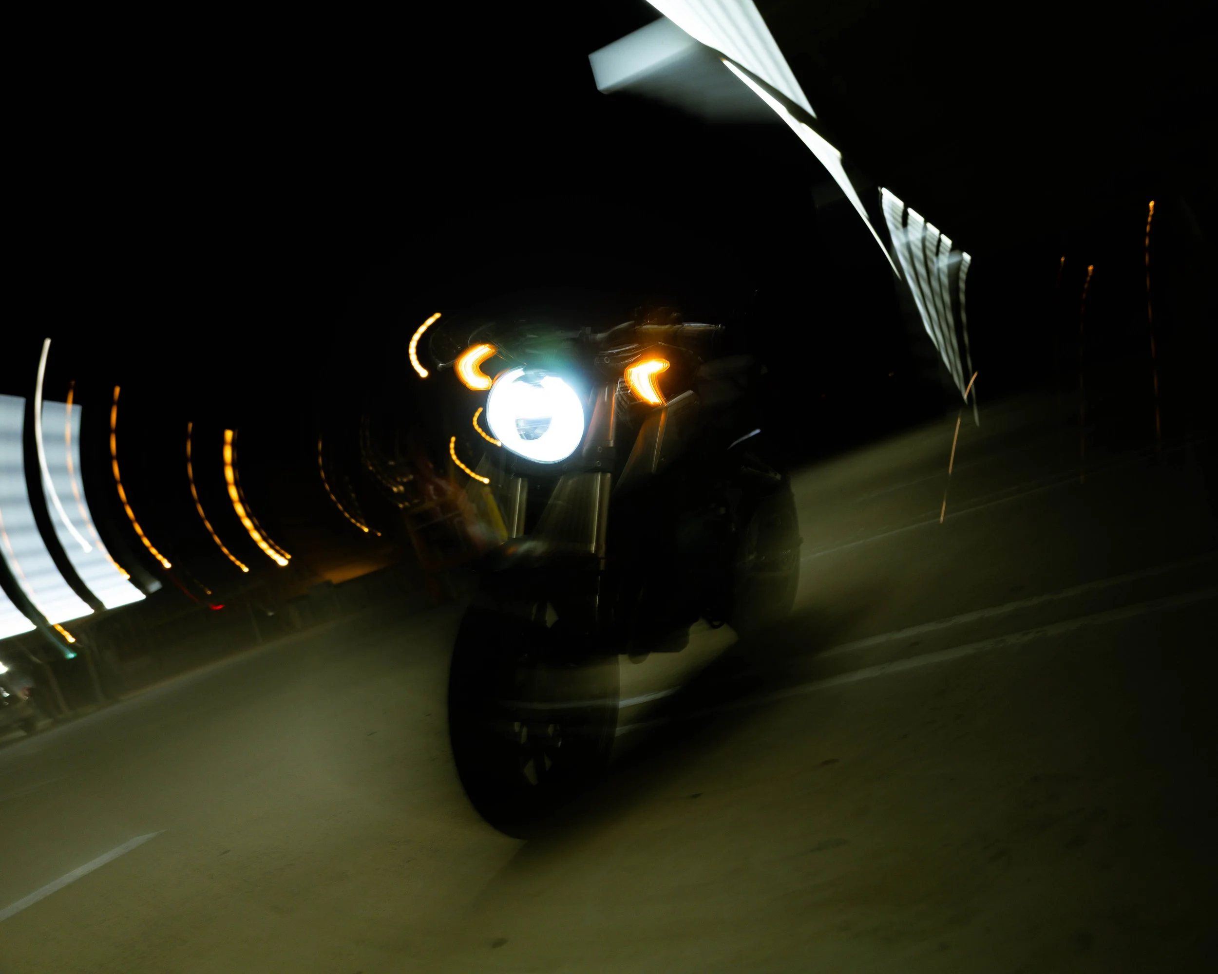 Nighttime scene of a motorcycle with its headlights on, leaning into a curve on a road with yellow and white light streaks from vehicle lights and reflections.
