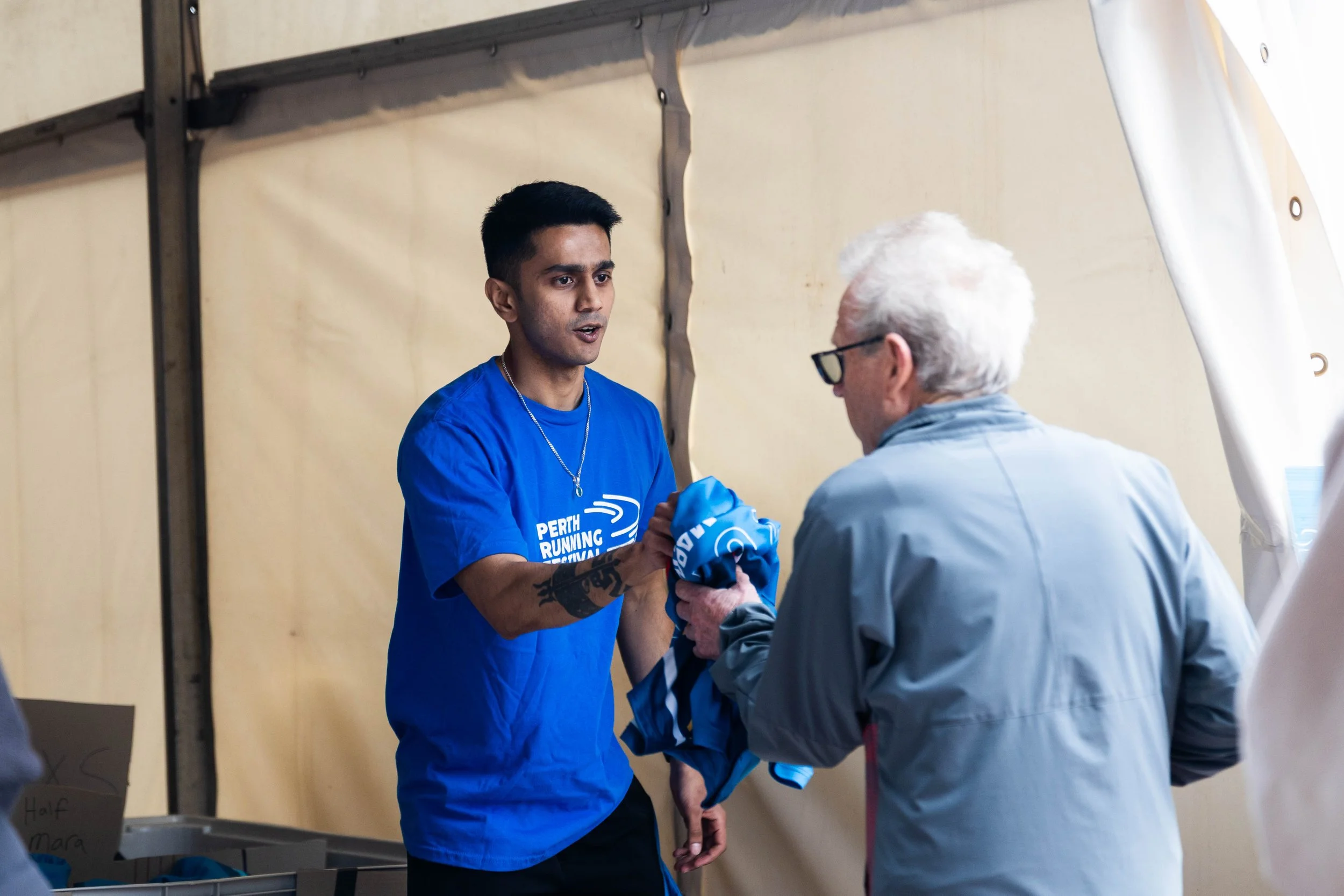 A young man wearing a blue shirt with the text "Perth Running Festival" is shaking hands with an older man wearing glasses and a gray jacket. They are in an indoor setting with a beige wall in the background.