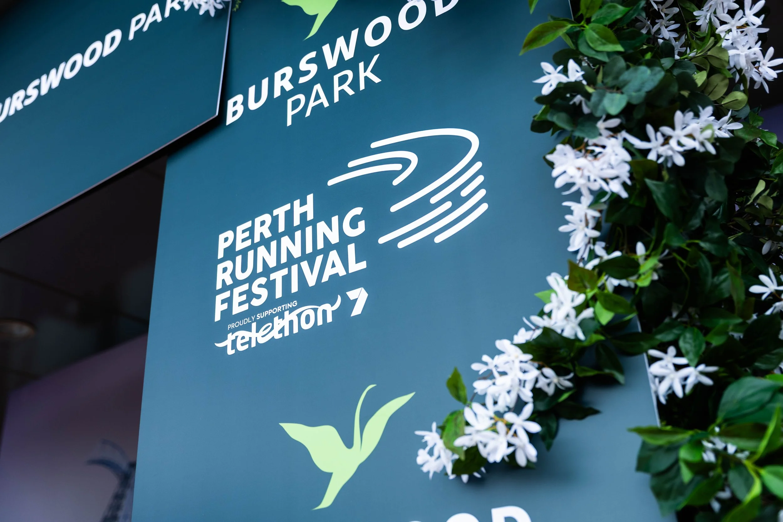 Sign for the Perth Burswood Park Running Festival, with white lettering on a blue background, surrounded by white flowers and green foliage.