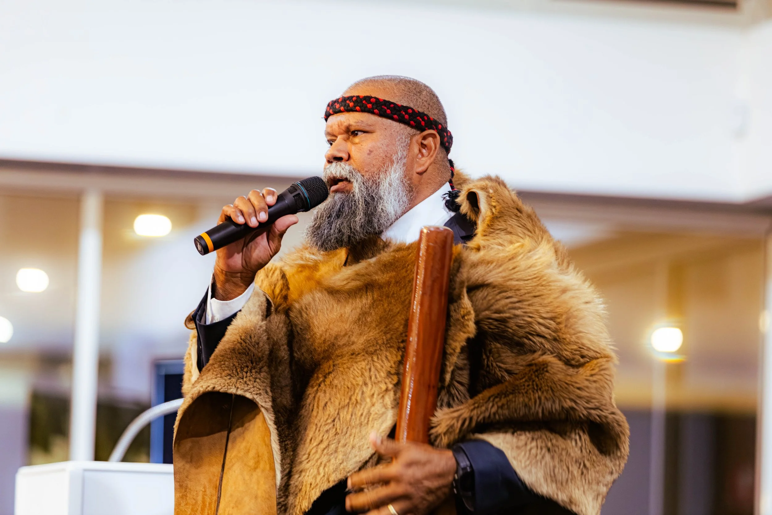 An elderly man with a beard, wearing a headband and a fur coat, holding a microphone and speaking.