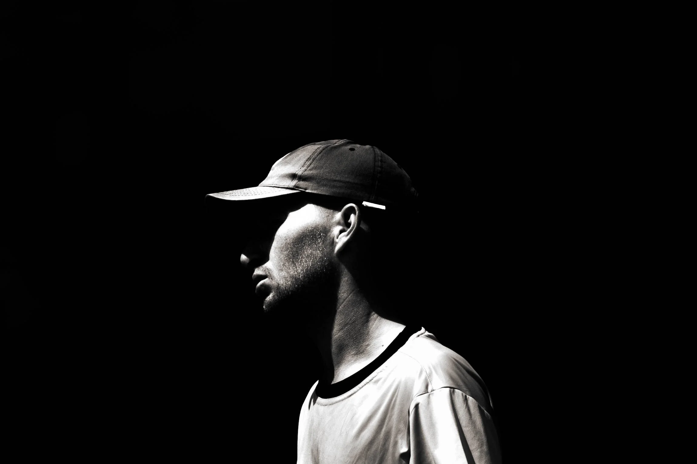 A person wearing a baseball cap and t-shirt, with their face turned to the side, in a high-contrast black and white photograph with a dark background.