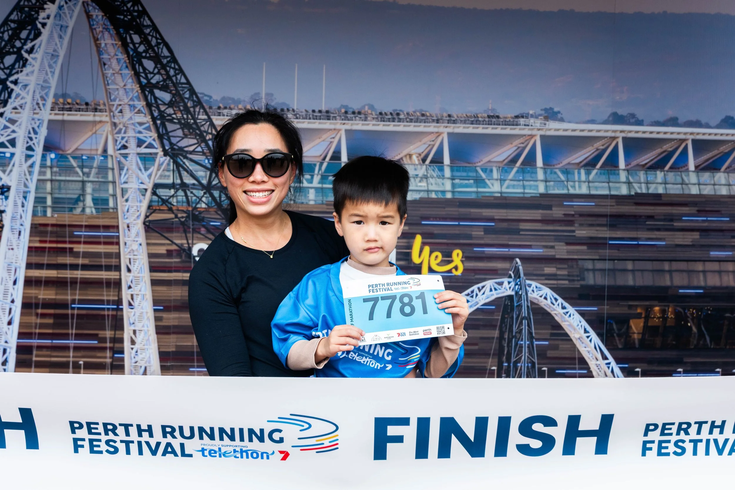 A woman and a young boy holding a marathon race bib at the finish line of the Perth Running Festival, with a Ferris wheel and cityscape in the background.