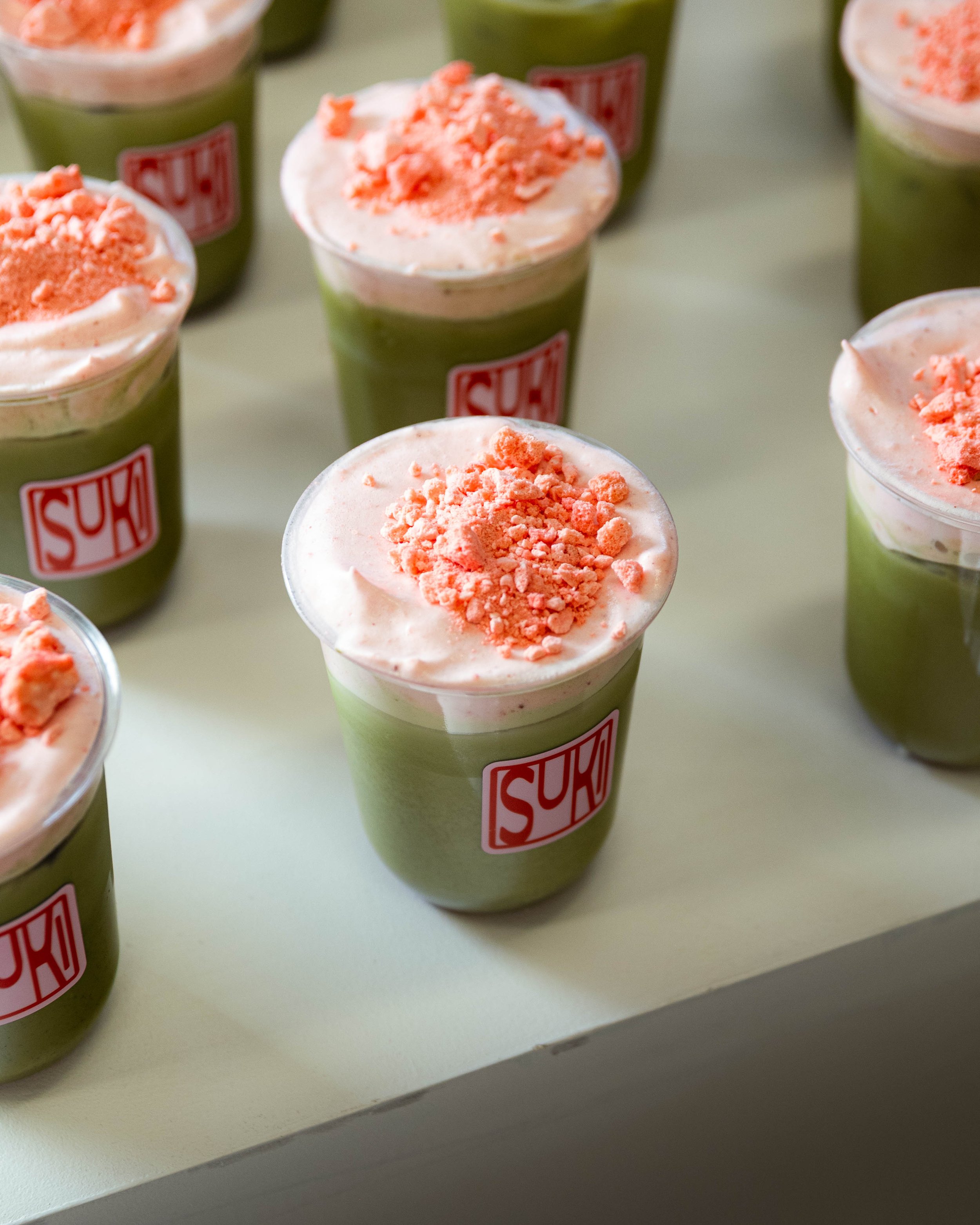 Several plastic cups filled with a green drink, topped with pink whipped cream and pink crumbles, placed on a white surface, with a red and white "SUK" logo on the cups.