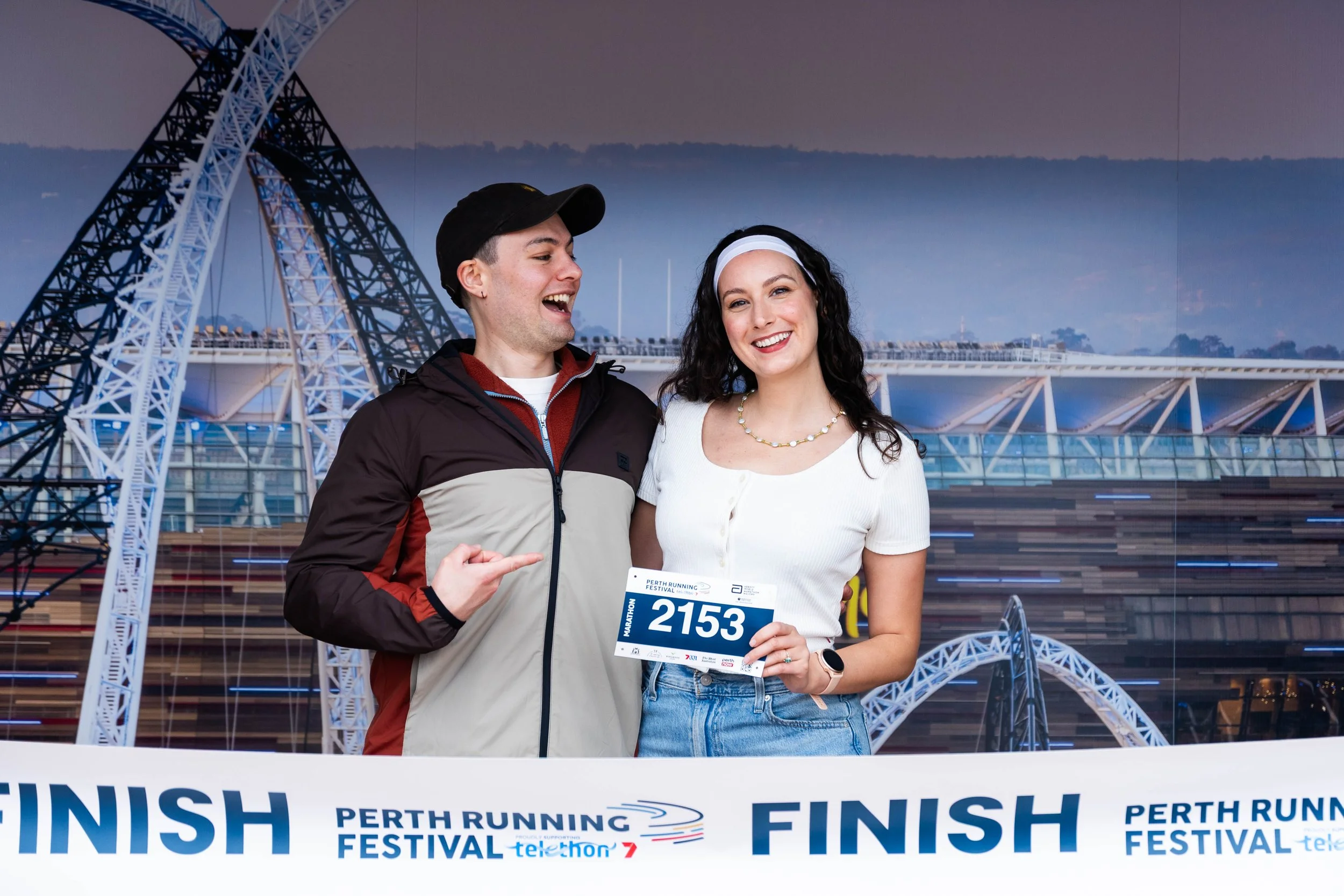 A man and a woman smiling at the camera after finishing a marathon, holding a race bib with the number 2153, standing in front of a cityscape with a large steel structure, under a cloudy sky.