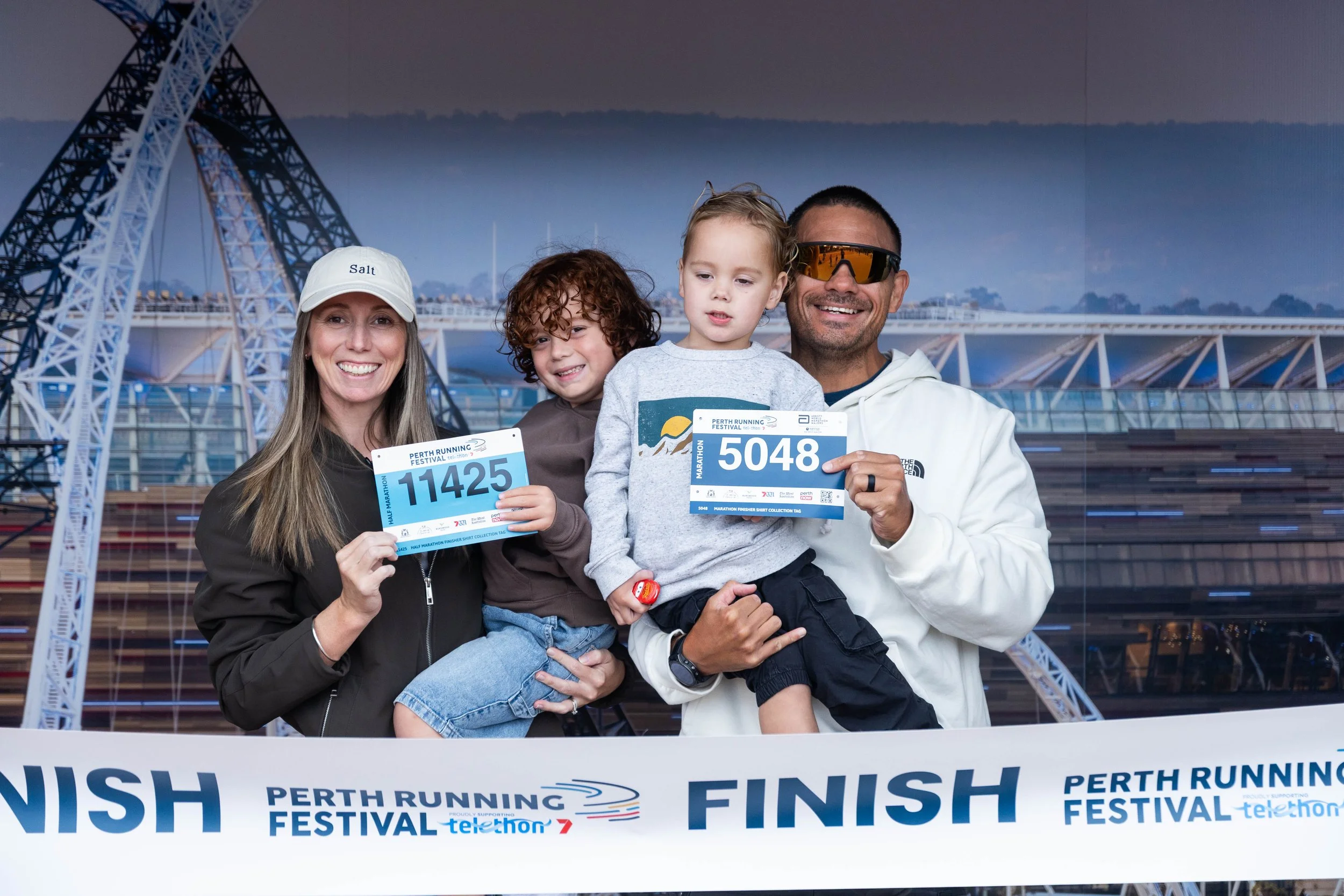 A family of four celebrates at the finish line of a marathon, holding race medals and bibs, with a bridge and cityscape in the background.