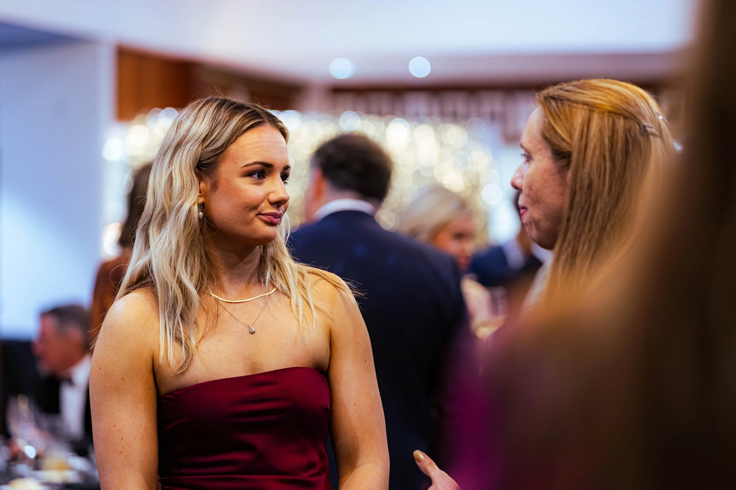 A young woman with long blonde hair, wearing a strapless maroon dress and jewelry, engaging in conversation at a social event. She is in focus with other people and festive lights blurred in the background.
