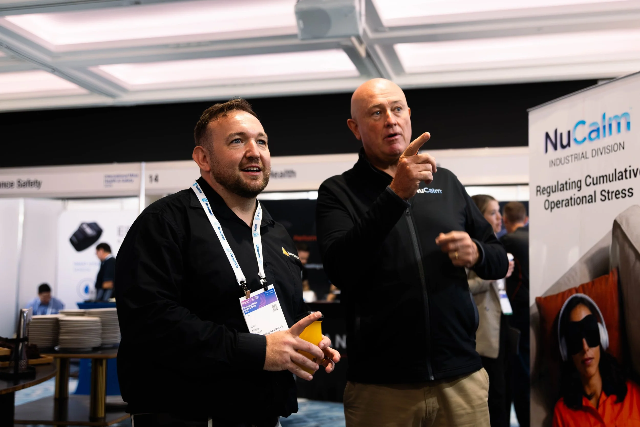 Two men at a conference booth, one holding a yellow cup with a yellow pen, engaging in conversation. The booth has a sign that reads 'NuCalm' and discusses regulating operational stress.