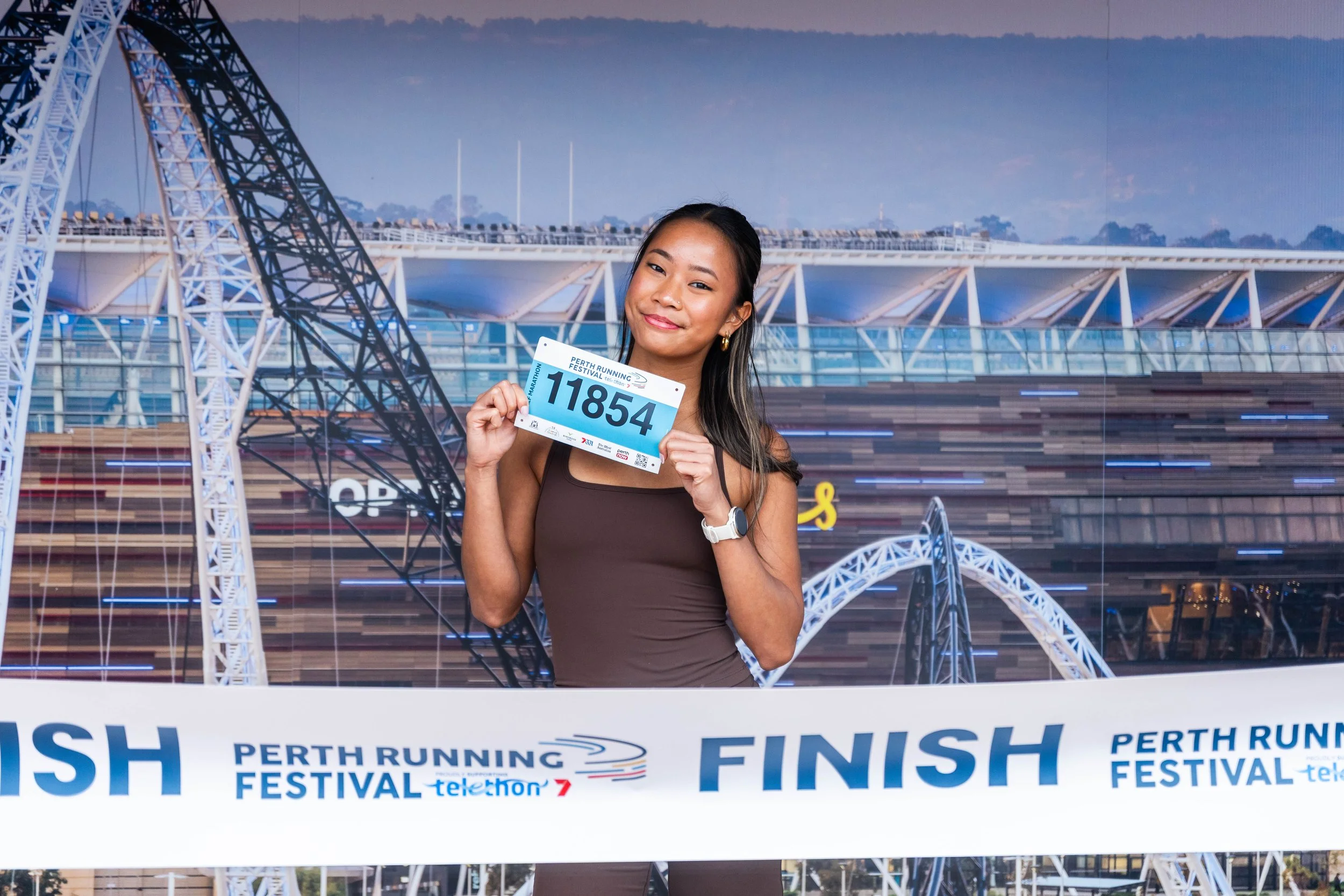 A young woman holding a bib with the number 11854 at the finish line of the Perth Running Festival, with a modern architectural structure in the background.