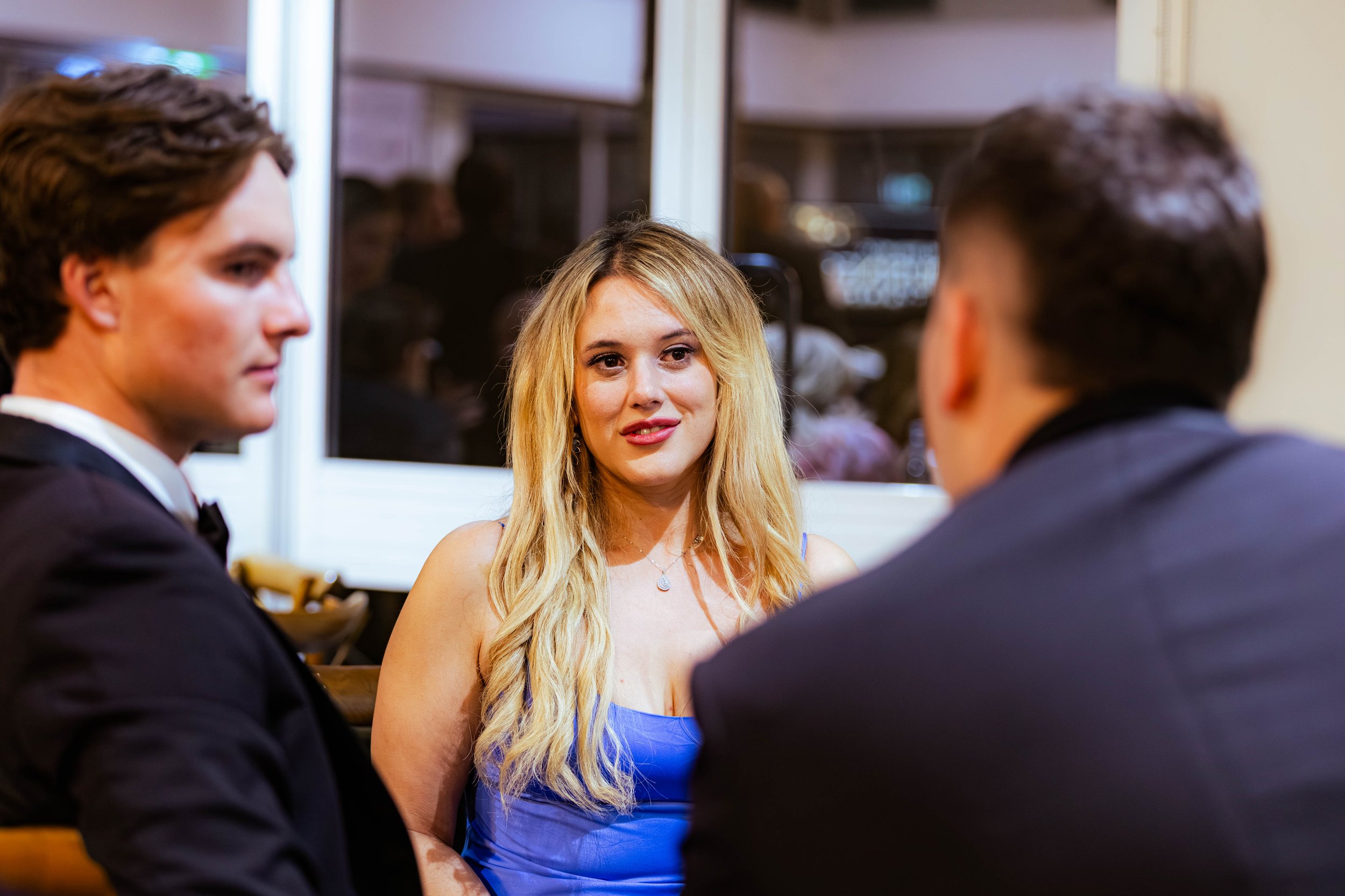 A woman with long blonde hair wearing a blue dress is conversing with two men in black tuxedos in a warmly lit indoor setting.