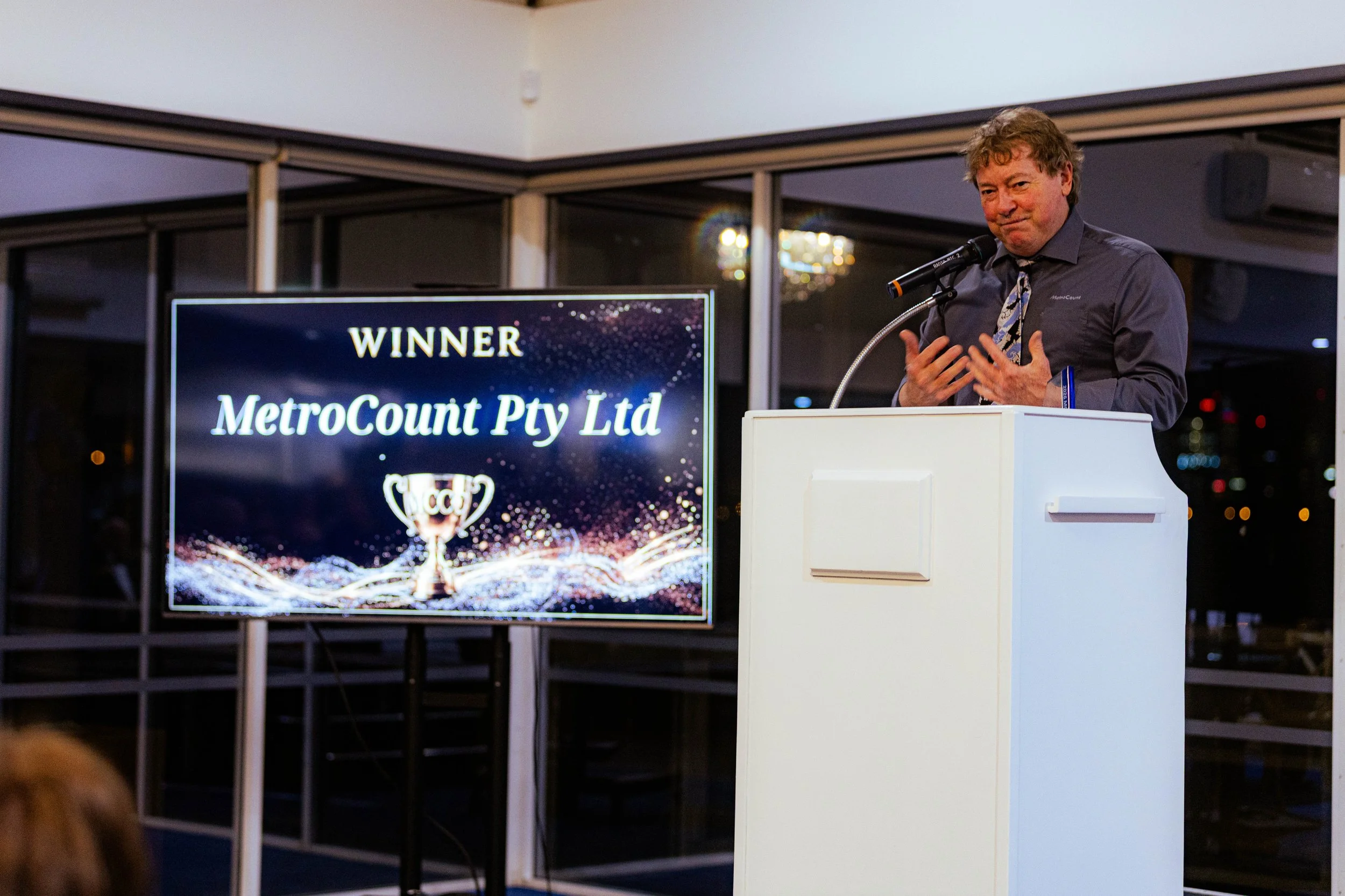 A man speaking at a podium during an award ceremony with a sign displaying 'WINNER MetroCount Pty Ltd' and a trophy graphic.