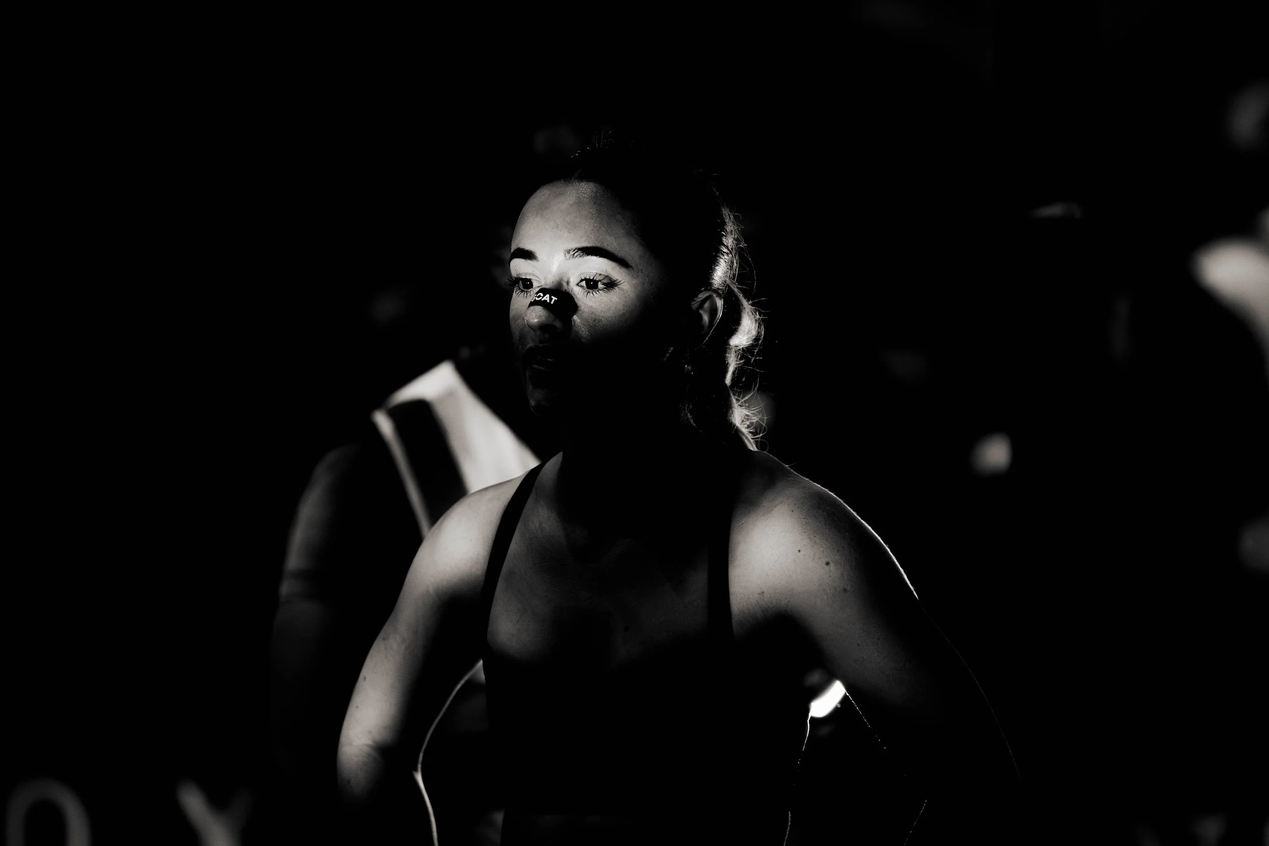 A woman with a taped nose, looking down, illuminated by a single light source in a dark setting.