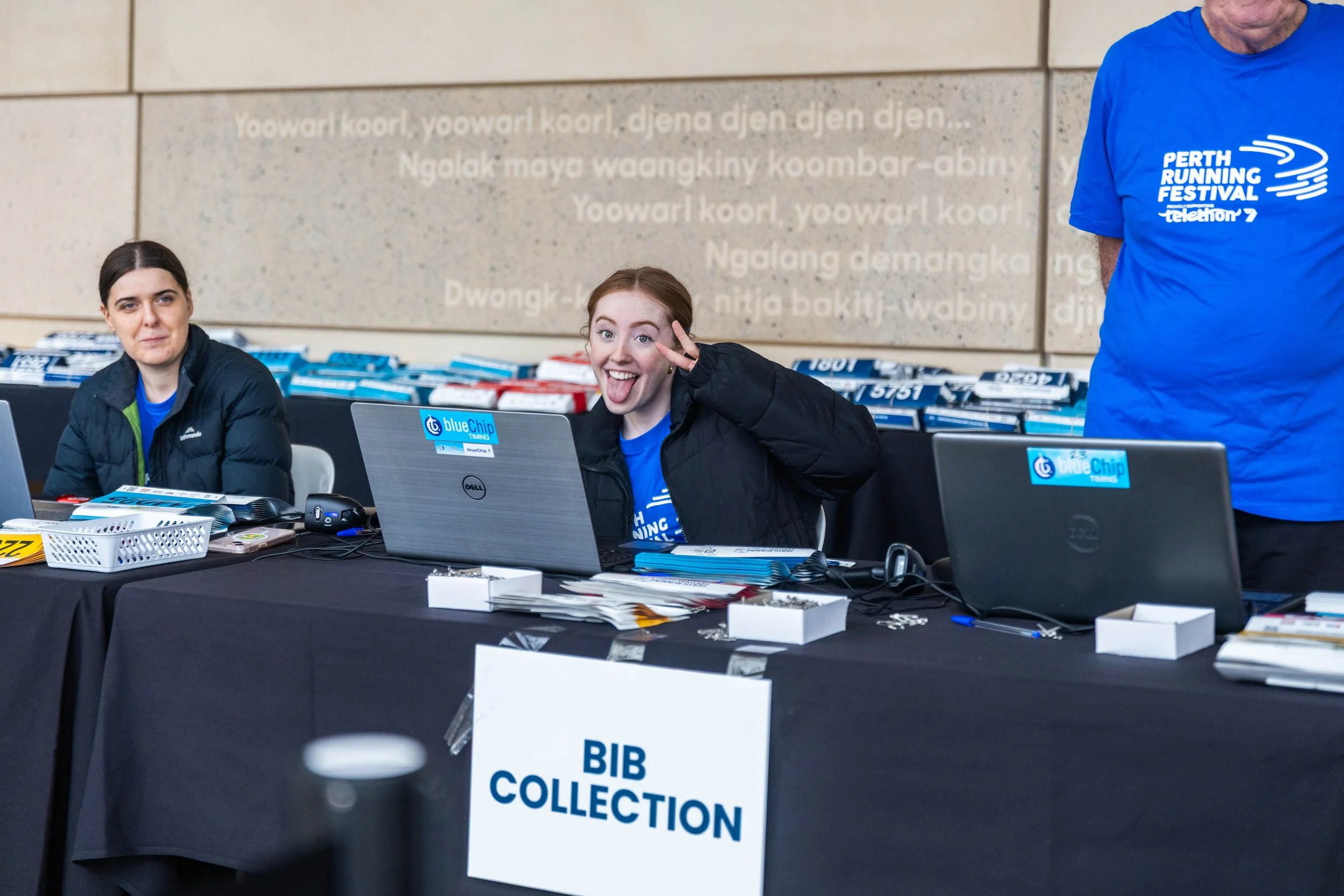 Three women seated at a table with laptops and paperwork at the Perth Running Festival volunteer registration. One woman is smiling and making a peace sign. The table has a sign reading 'Bib Collection.' A man in a blue shirt stands to the right. The
