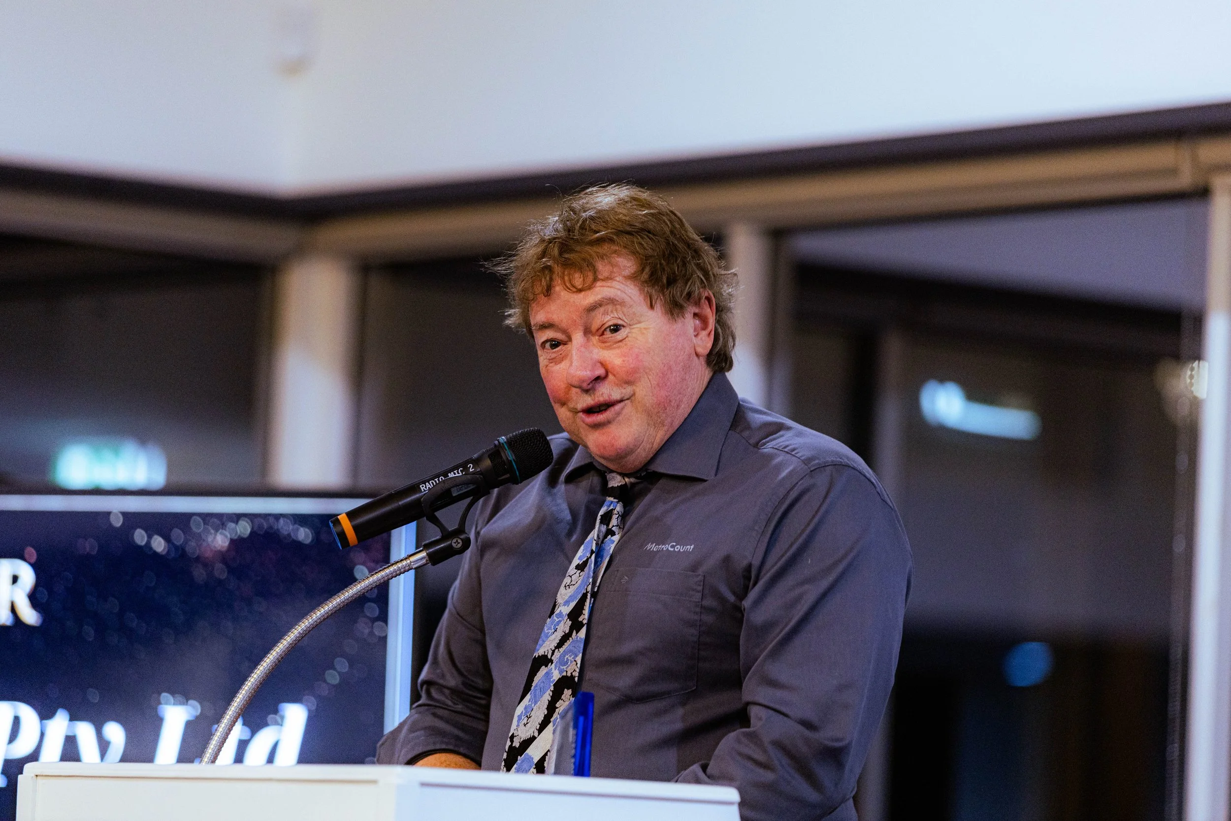 Man in gray shirt with tie speaking into microphone at a podium, with a screen in the background displaying text and a starry background.