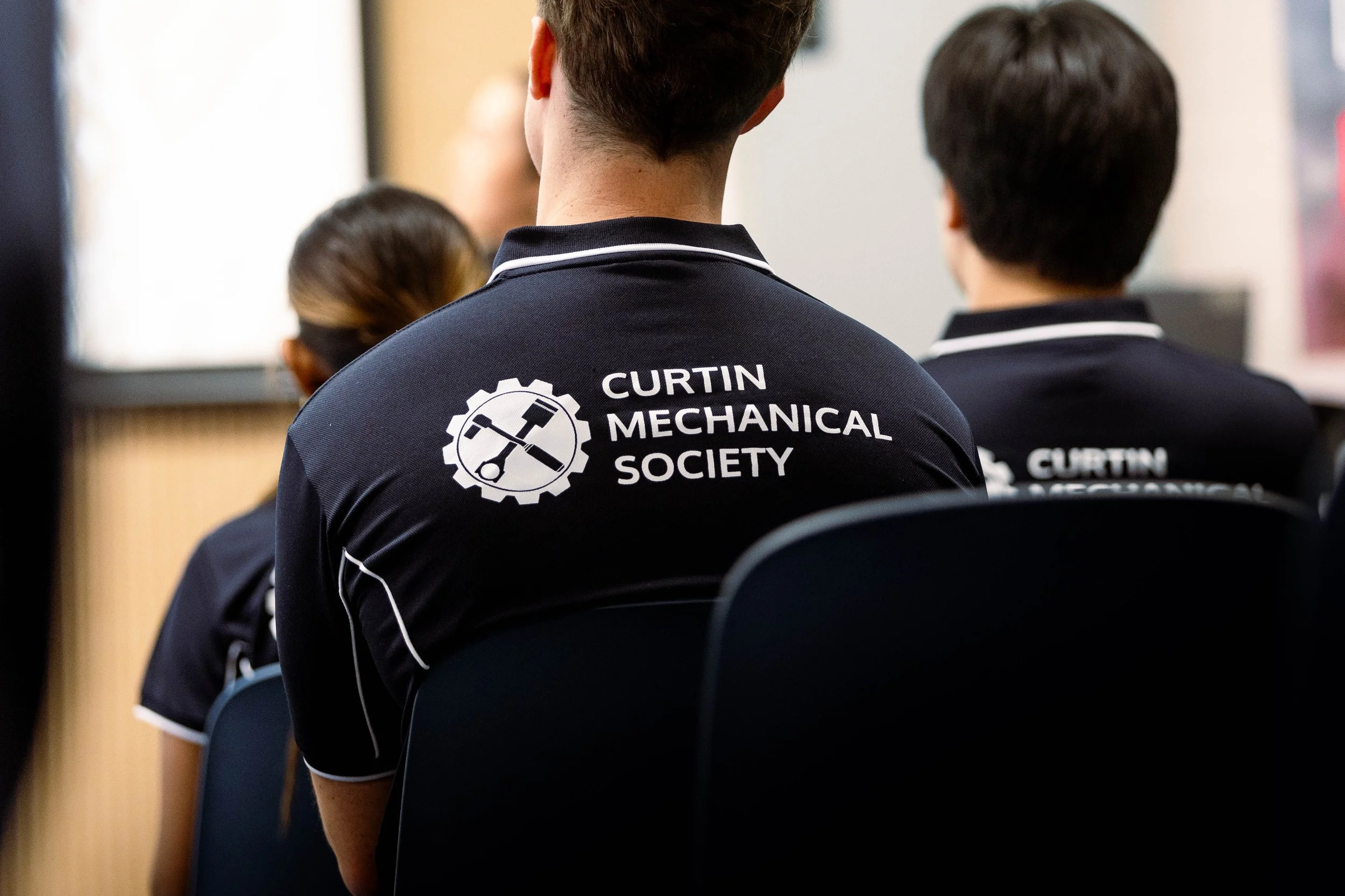 People attending a meeting or conference, with one person wearing a navy blue polo shirt with the logo and text 'Curtin Mechanical Society' on the back. The participants are seated facing forward, likely listening to a presentation or speaker.