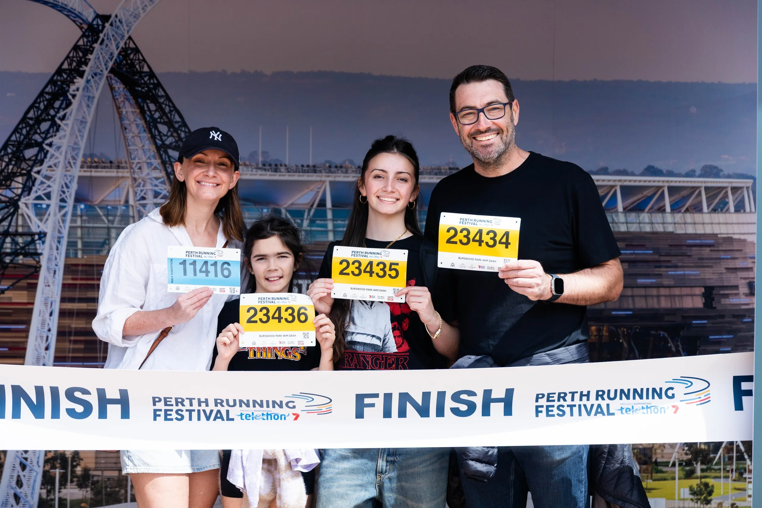 A family of five celebrating at the finish line of a running event, holding race bibs, with a modern bridge and cityscape in the background.