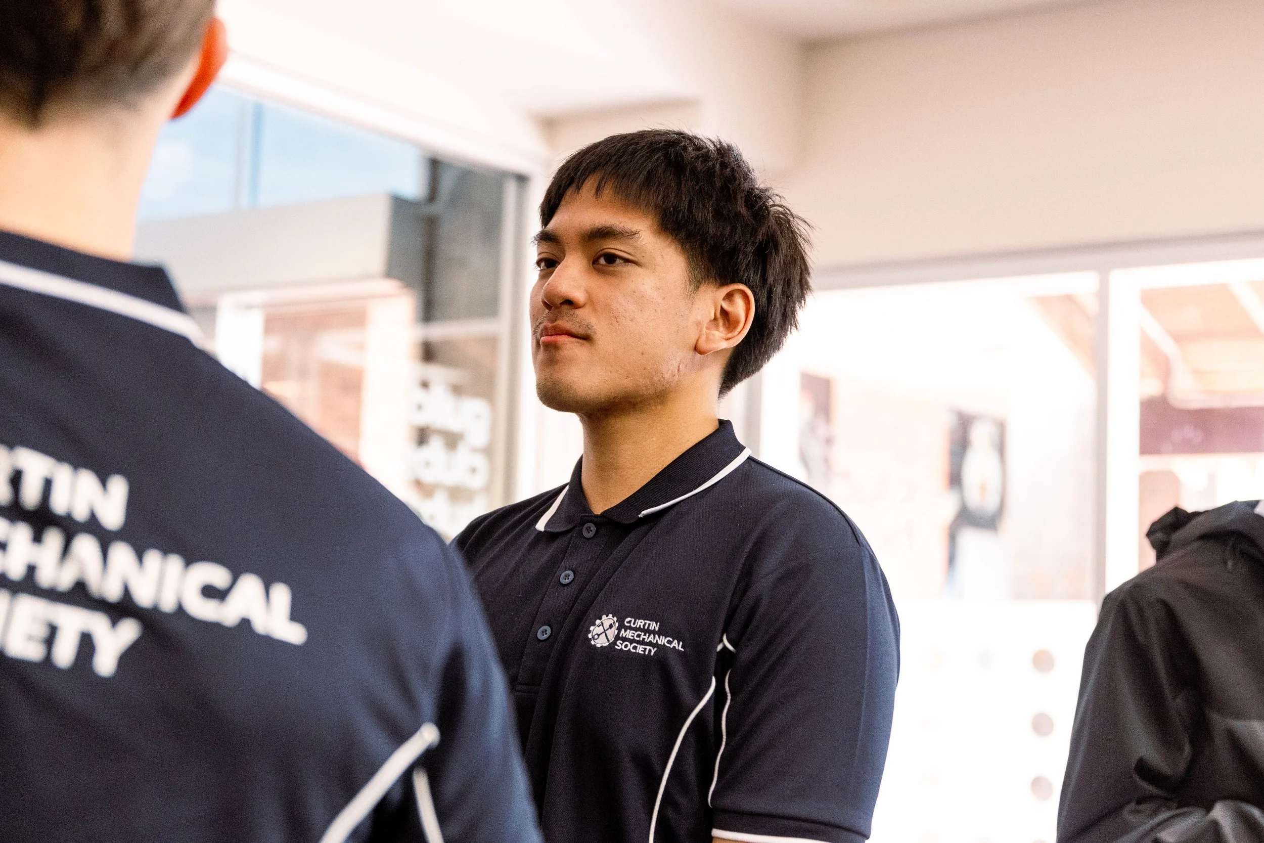 Young man wearing a black polo shirt with school logo, engaging in conversation with others, in a bright indoor setting.