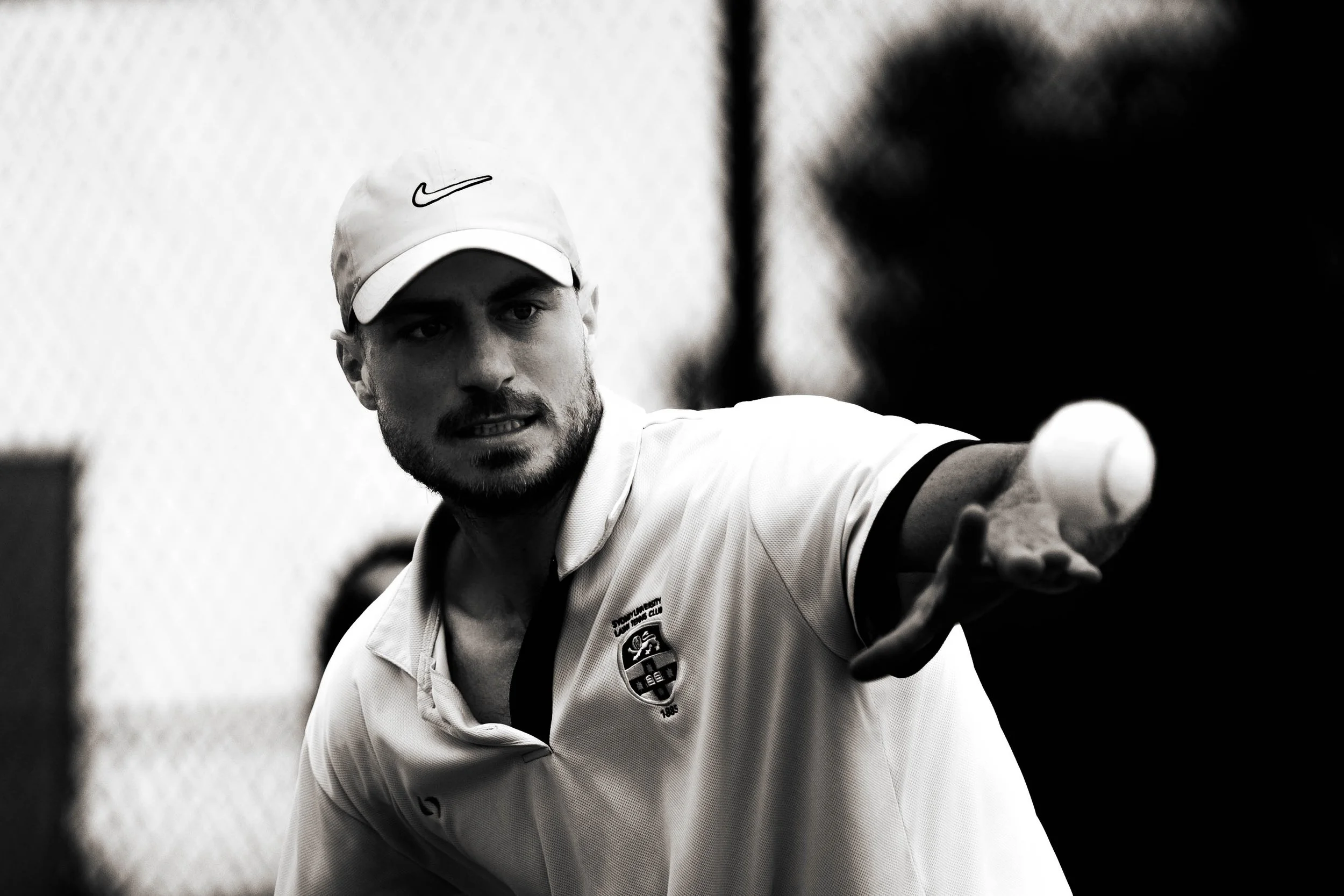 A man with a beard wearing a white sports shirt and a white cap, possibly a tennis player, reaching out with his hand.