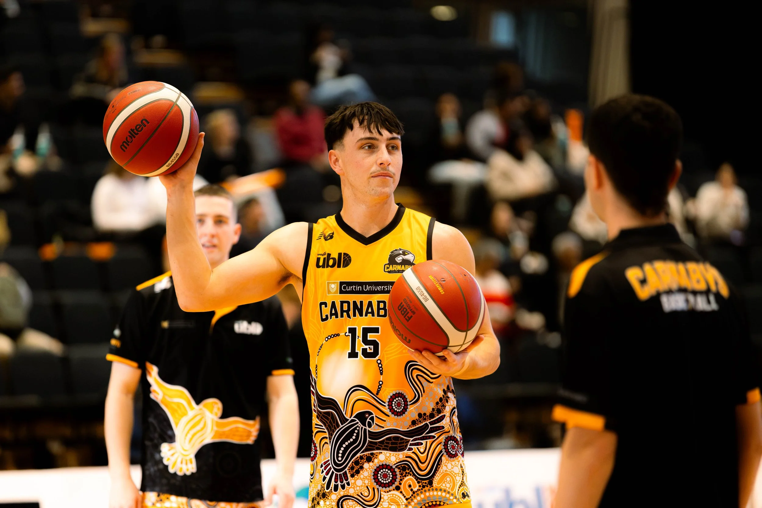 A basketball player wearing a yellow jersey with the number 15, holding a basketball with his right hand, and standing on a basketball court. The jersey has the words 'CARNABY' and a logo, with a patterned design. There are other players and spectato