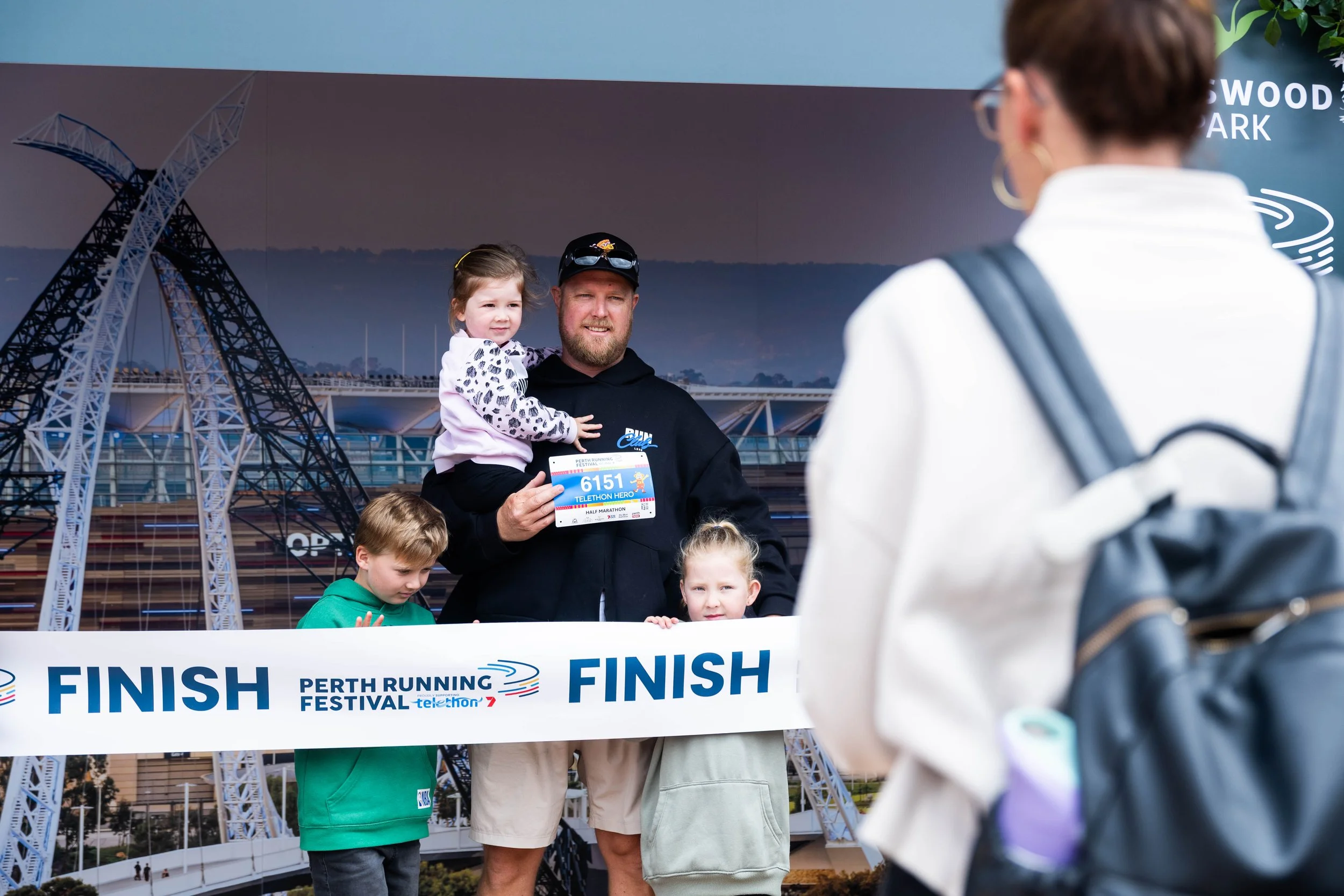 A man holding a young girl at a finish line of a running event, with other children around, and a woman in the foreground. The event is the Perth Running Festival, with a large banner reading 'FINISH' and the event logo.
