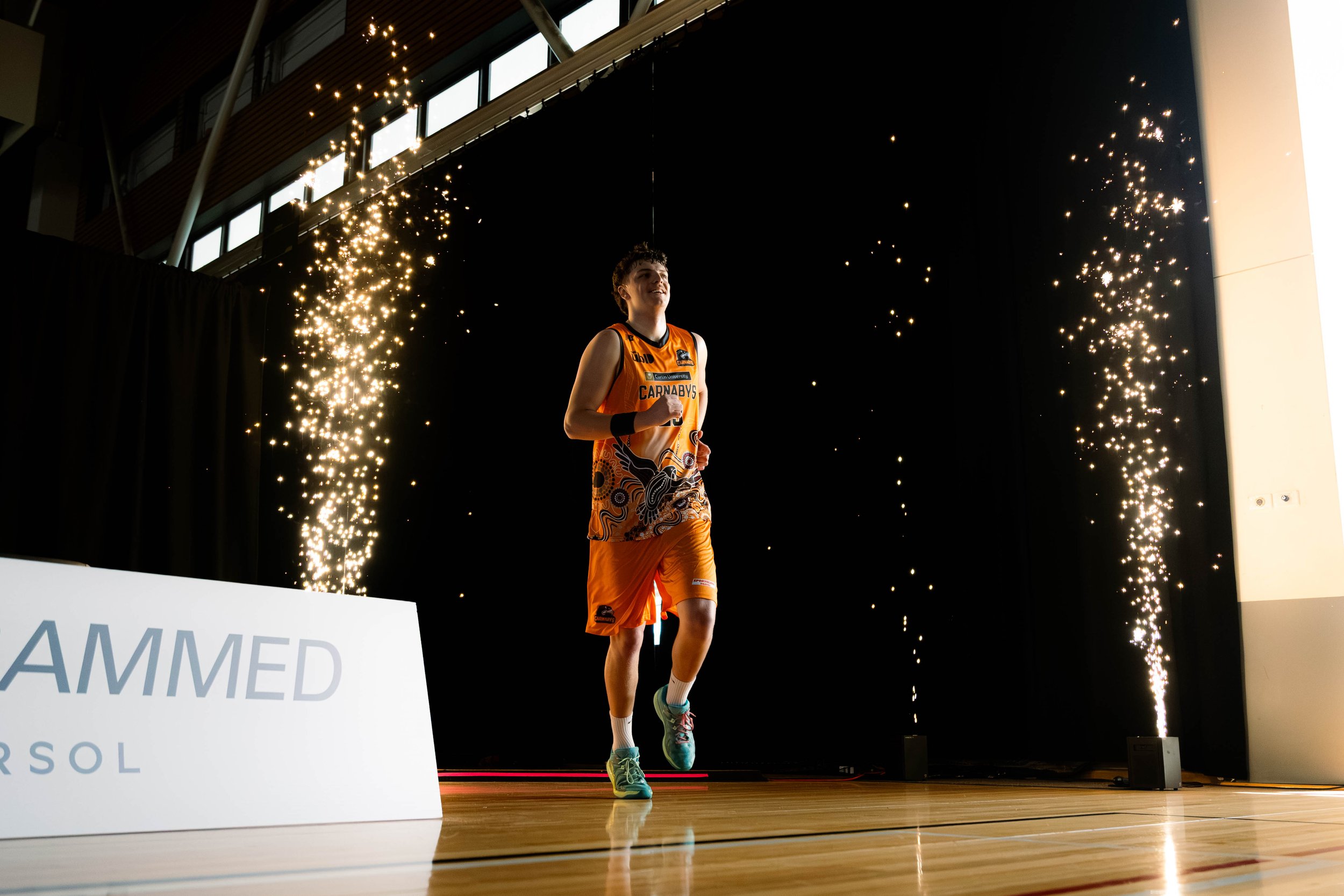 A young male athlete in an orange sports uniform running on a stage with sparks flying around him during an indoor event.