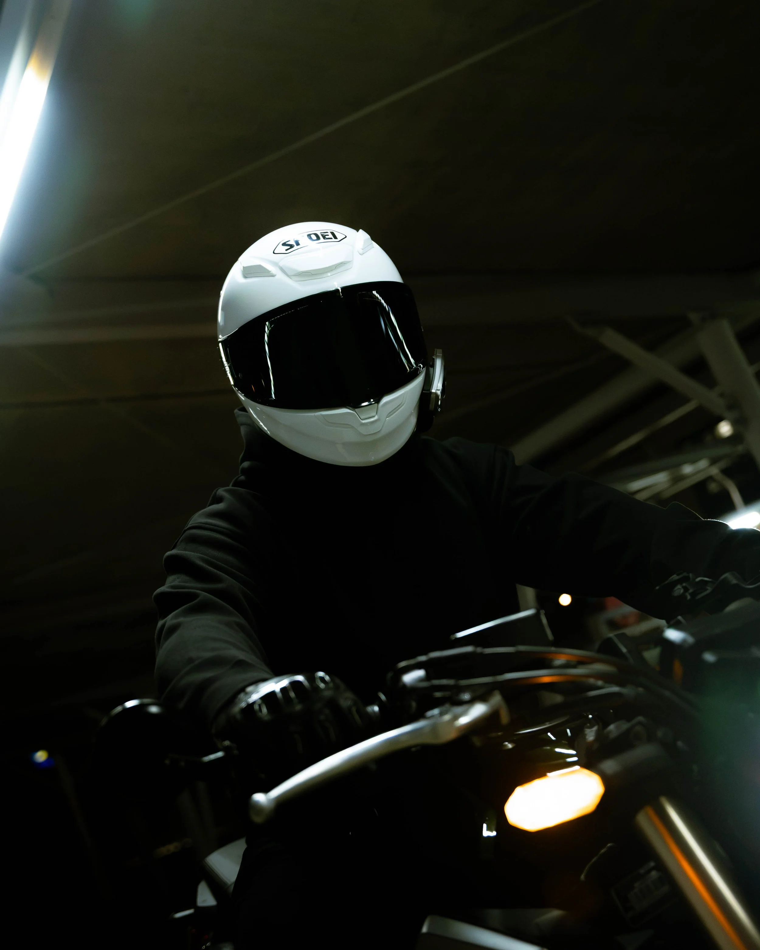 Person wearing a white motorcycle helmet with a black visor, taking a selfie on a motorcycle in a dimly lit underground parking garage.
