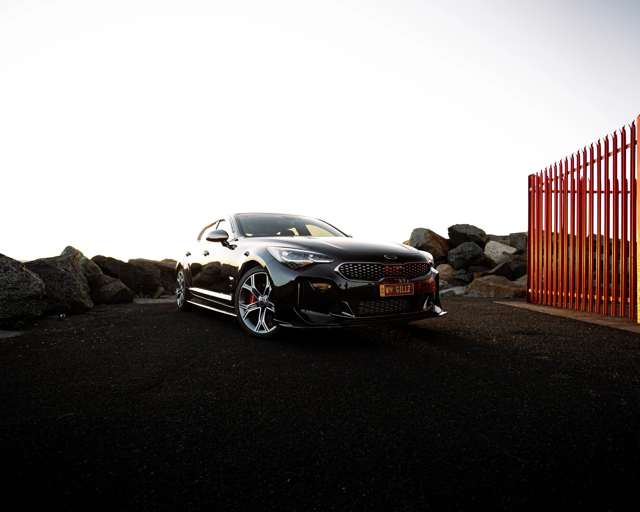 A black sports car parked on a rocky surface next to a red metal fence, with large rocks scattered around, and a clear bright sky overhead.