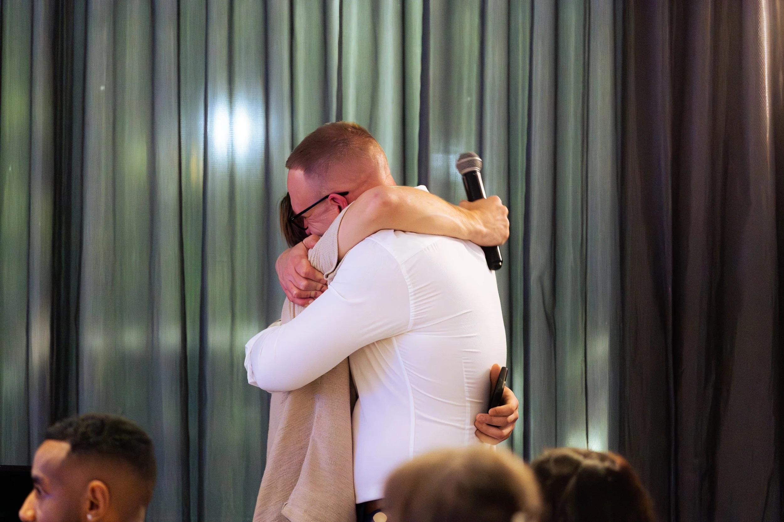 A man and a woman are hugging each other tightly, with the man holding a microphone in his right hand. The background features cloudy green and black curtains, suggesting a formal or staged event.