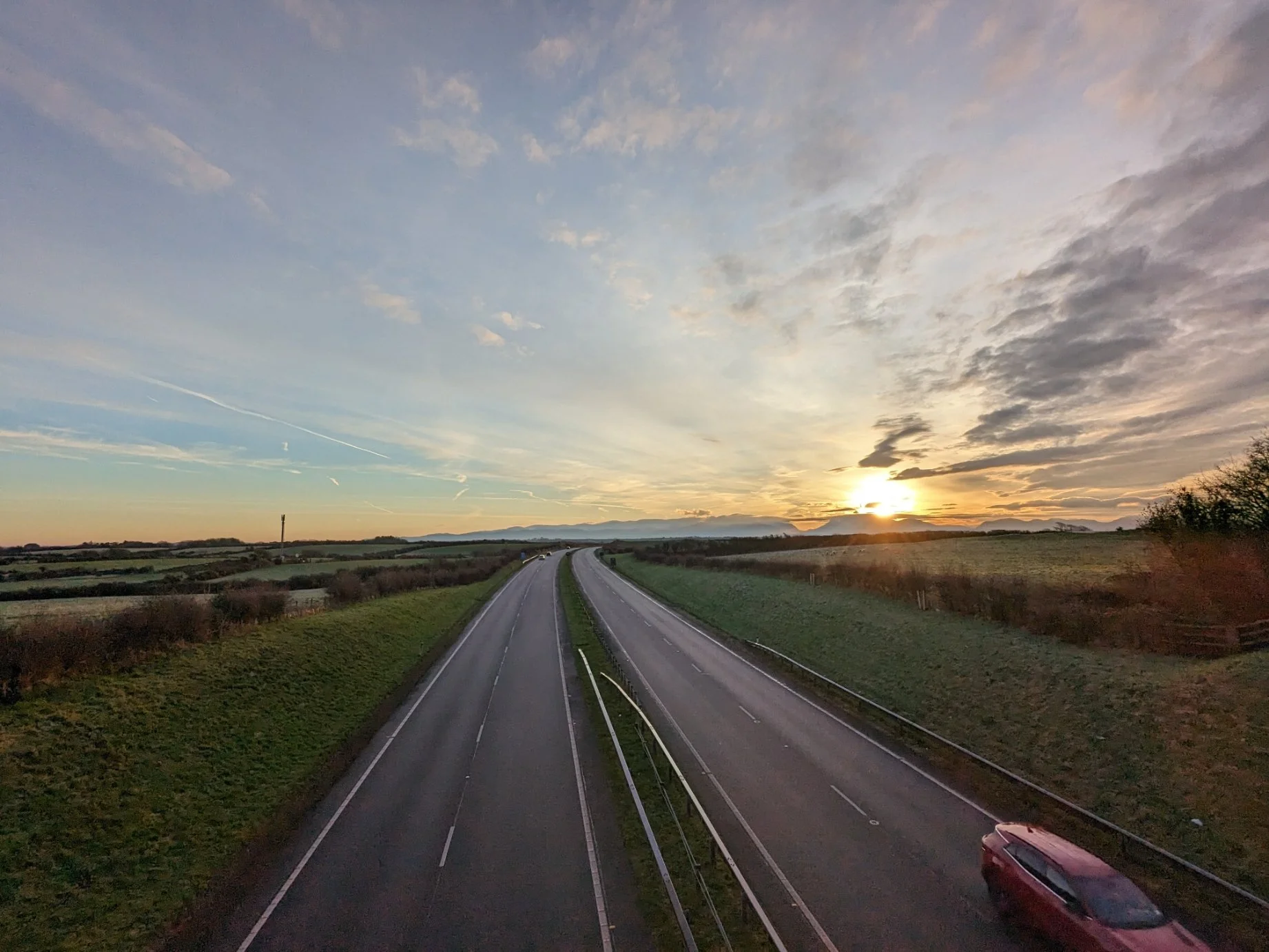 A rural highway at sunset with a car in the lower right corner, green fields on either side, and a sky with clouds and the sun near the horizon.