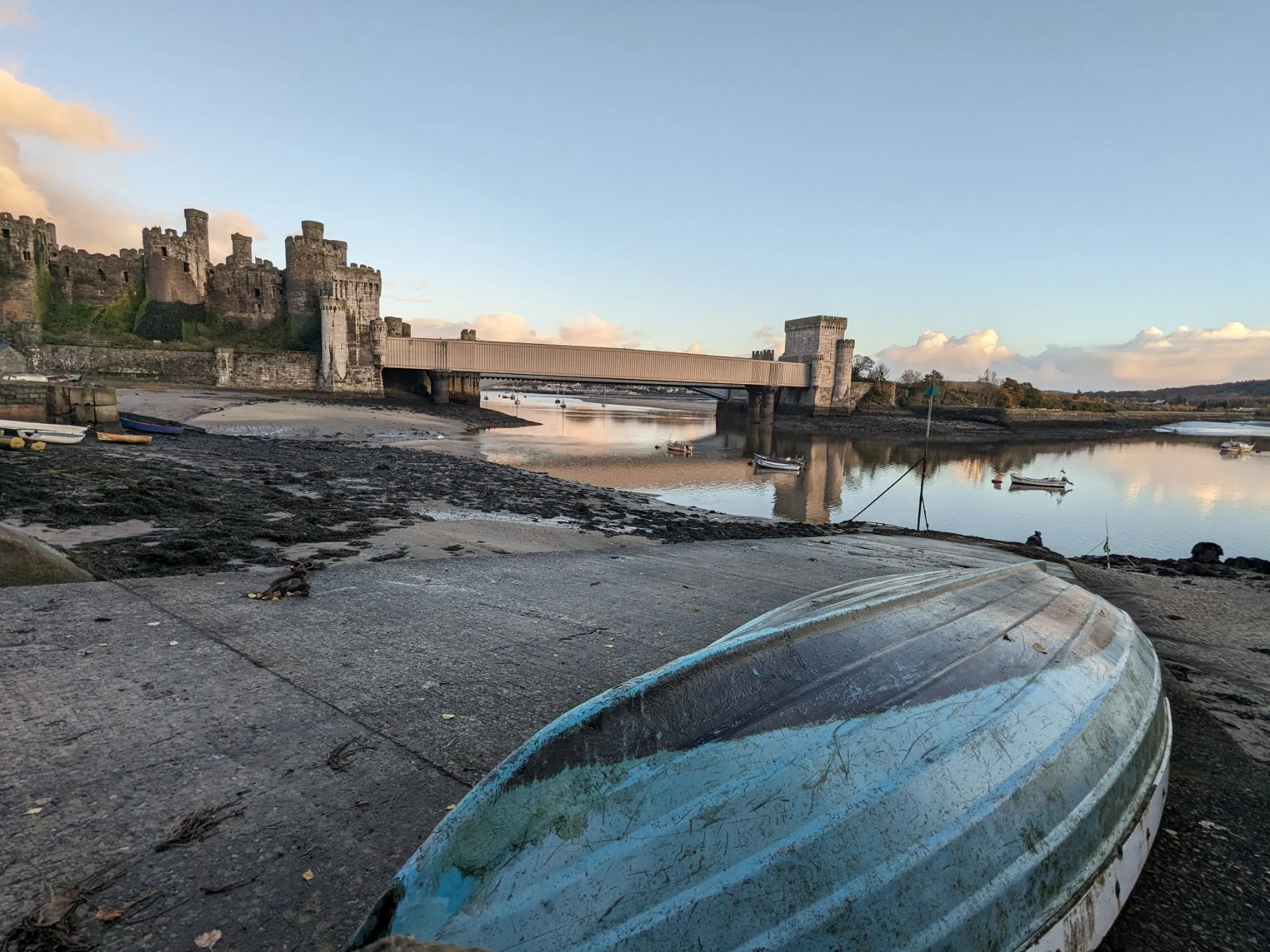 A scenic view of a historic castle with stone walls and towers located near a calm river, with boats anchored on the water and a bridge connecting the castle to the land. The foreground shows a weathered boat and a sandy, rocky shoreline.
