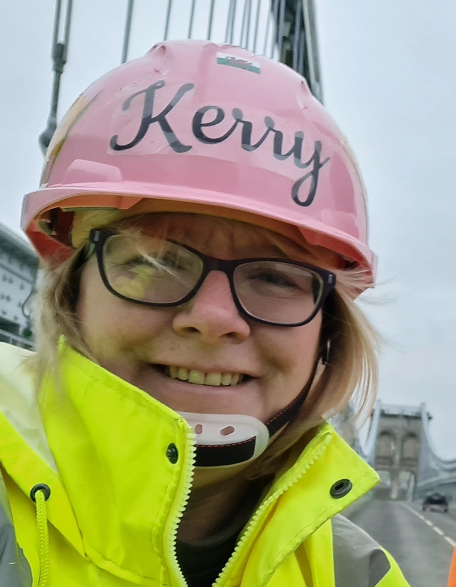 A person wearing a pink safety helmet with 'Kerry' written on it, glasses, and a yellow high-visibility jacket, smiling at a construction site.