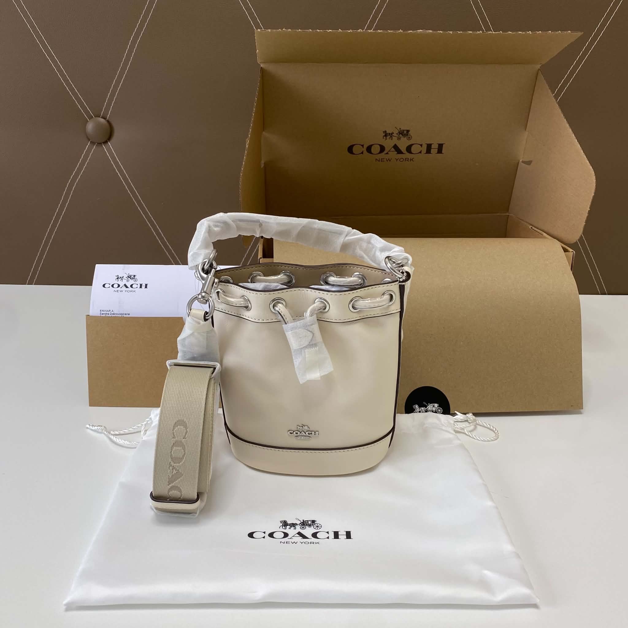 Bolso de piel beige con cadena gruesa plata, en una caja de cartón con la marca Coach de color negro. La caja tiene la inscripción 'Coach New York' con el logo de un carruaje de caballos.