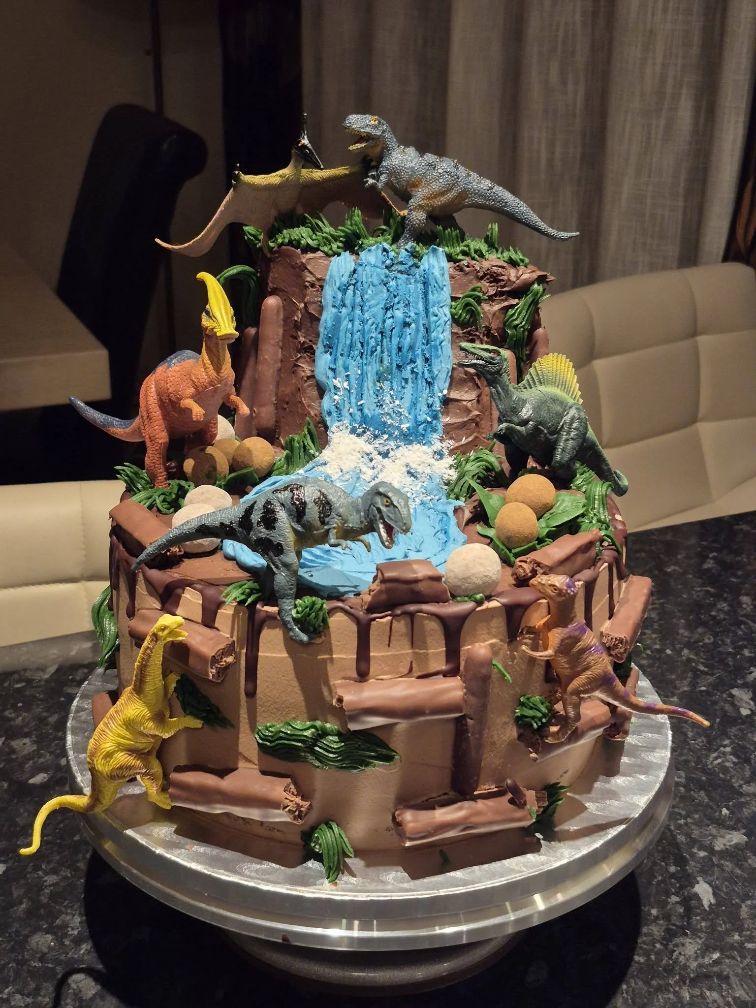 2-layer jurrasic park inspired birthday cake decorated with various sweets, icing and toy dinosaurs.