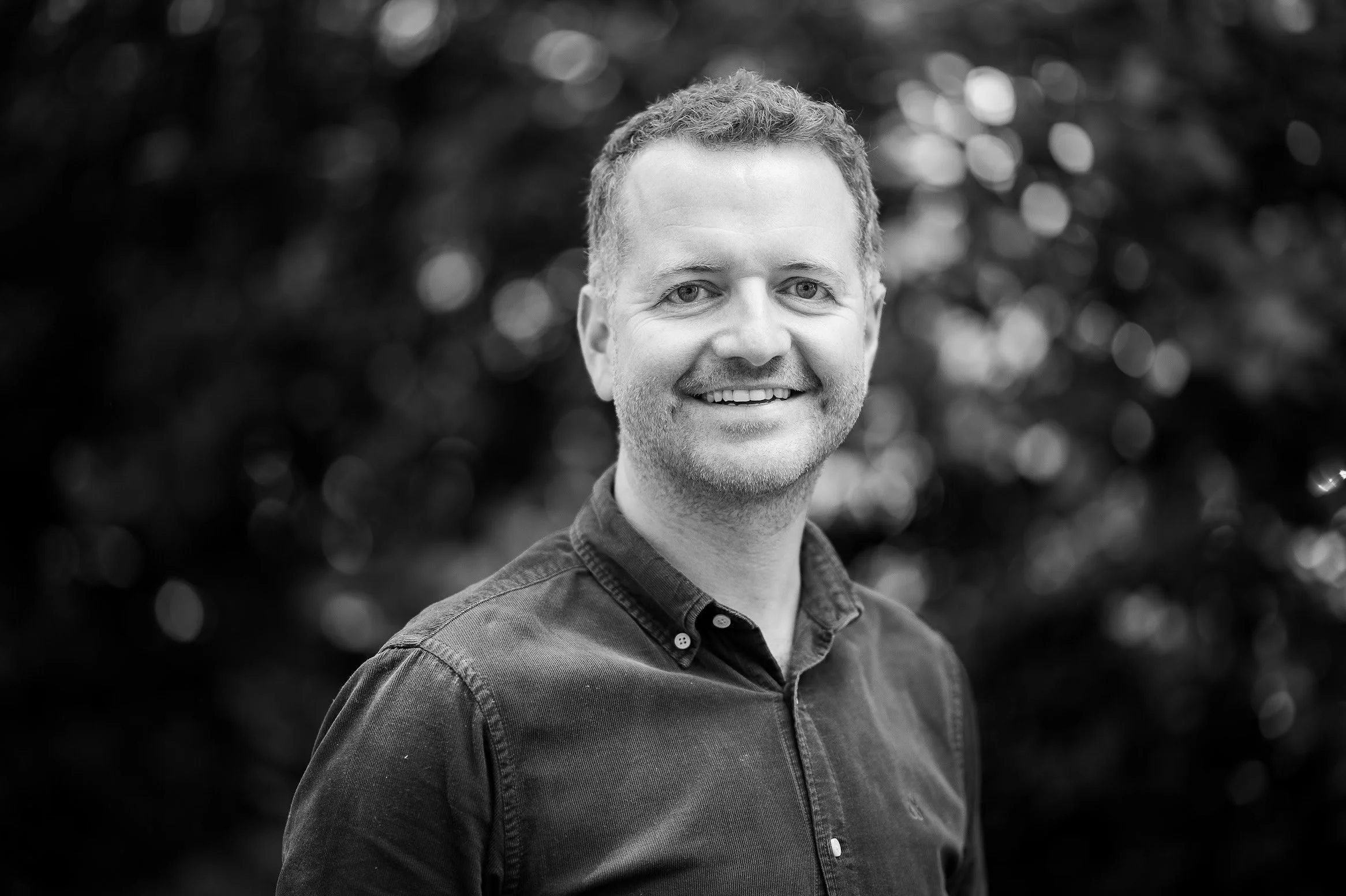 Black and white portrait of a smiling man with short curly hair, wearing a collared shirt. Background is blurred with bokeh effects.