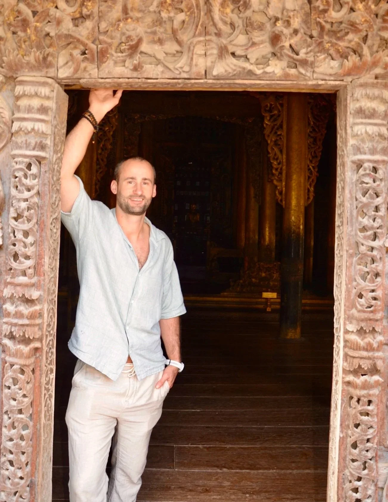 A man with short hair, beard, and light-colored shirt posing inside an intricately carved wooden doorway, with a dark ornate interior behind him.