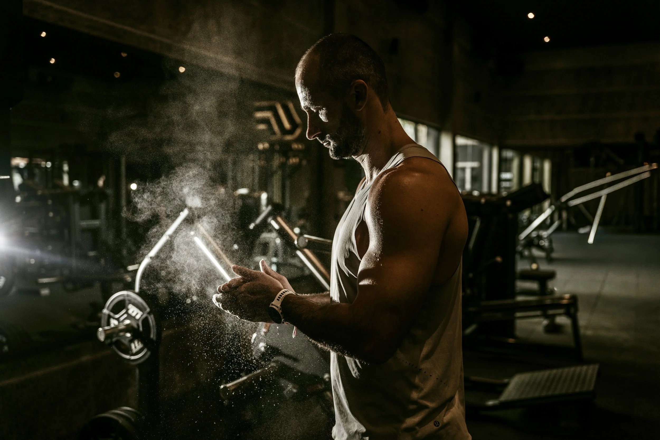 A man with short hair and a beard is in a gym, looking down and touching his hands, with a cloud of chalk dust around them. The gym has exercise equipment and large windows in the background.