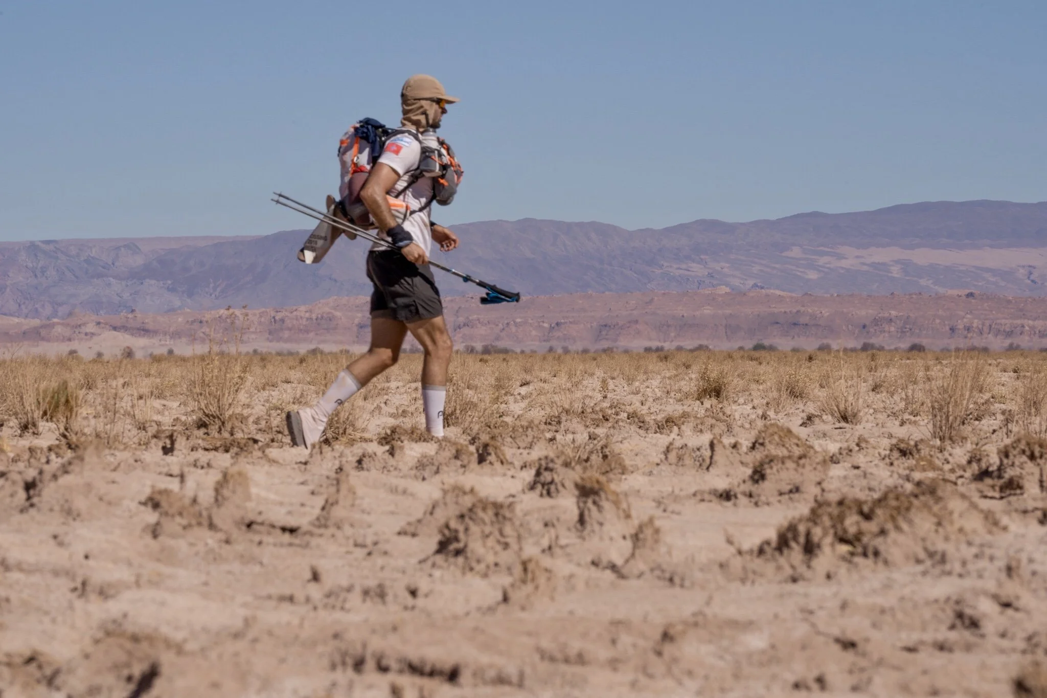 athletic running, ultramarathon,  in a desert landscape with mountains in the background, carrying trekking poles and a backpack.