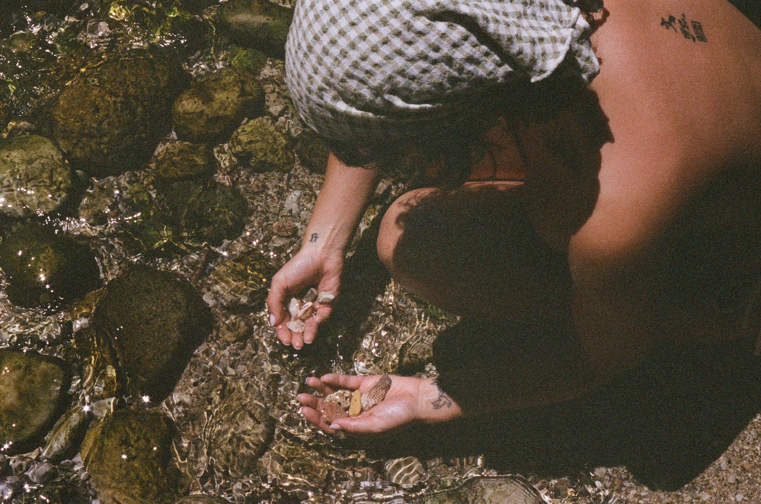 Person with a headscarf, reaching into water with rocks, collecting shells or small objects.