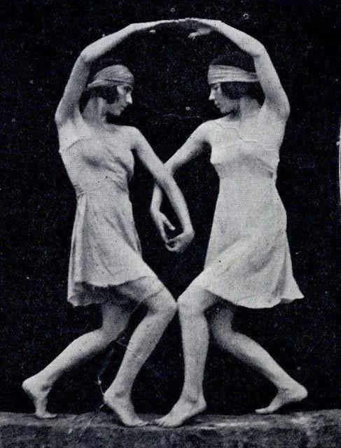 Two women wearing short dresses and headbands, holding hands, dancing or performing a pose against a dark background.