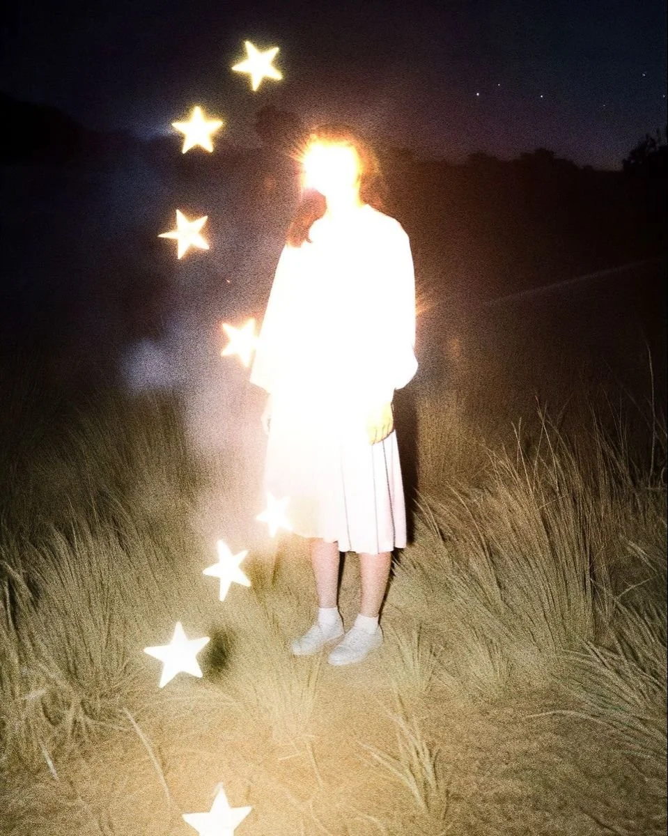 Person standing outdoors at night, with bright light obscuring their face, surrounded by glowing star-shaped effects and a dark sky with a few visible stars.