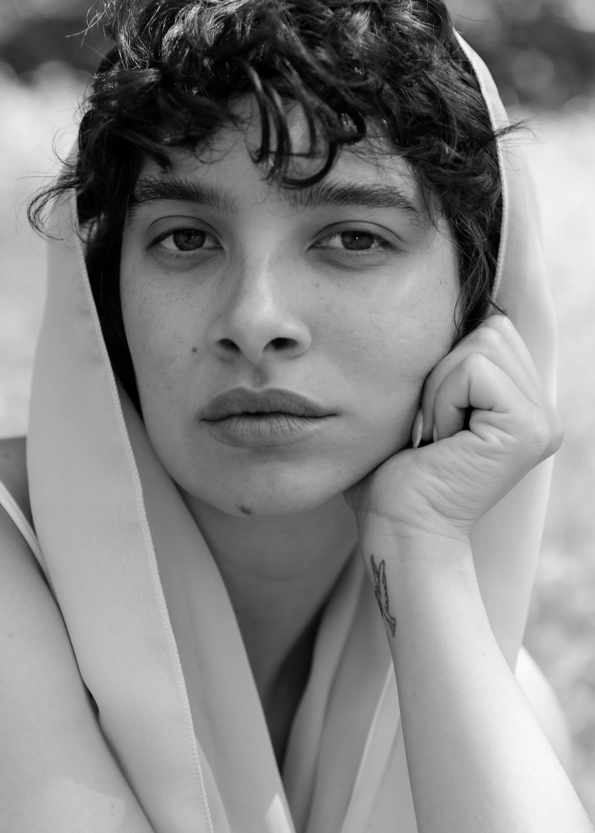 A black and white close-up portrait of a young person with curly hair, resting their face on their hand, looking directly at the camera.