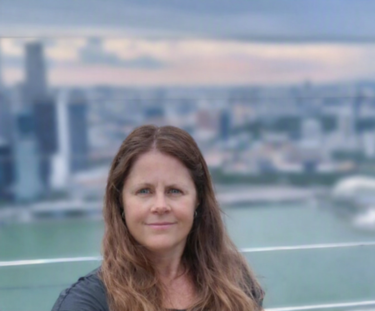 A woman with long brown wavy hair standing outdoors with a city skyline and bridge in the background.