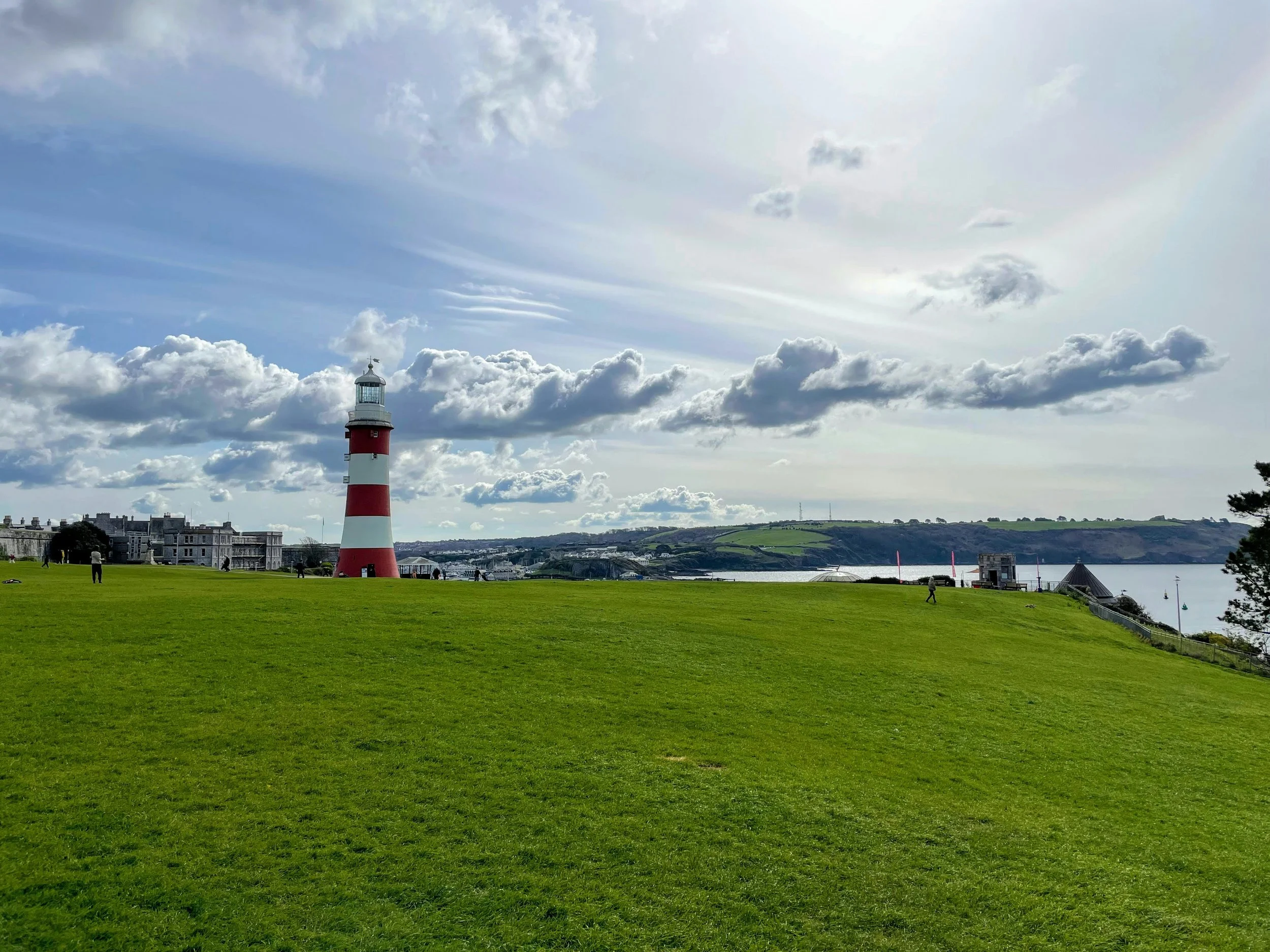 A seaside scene with a tall red and white striped lighthouse near the water, a grassy park with people walking, and a backdrop of hills and cloudy sky.
