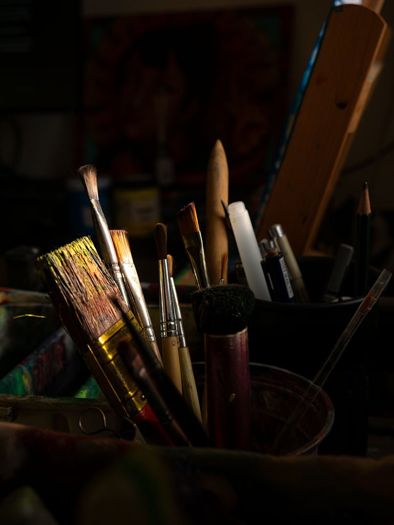 A collection of paintbrushes and art tools in a container on an artist's workspace, with a wooden easel in the background.