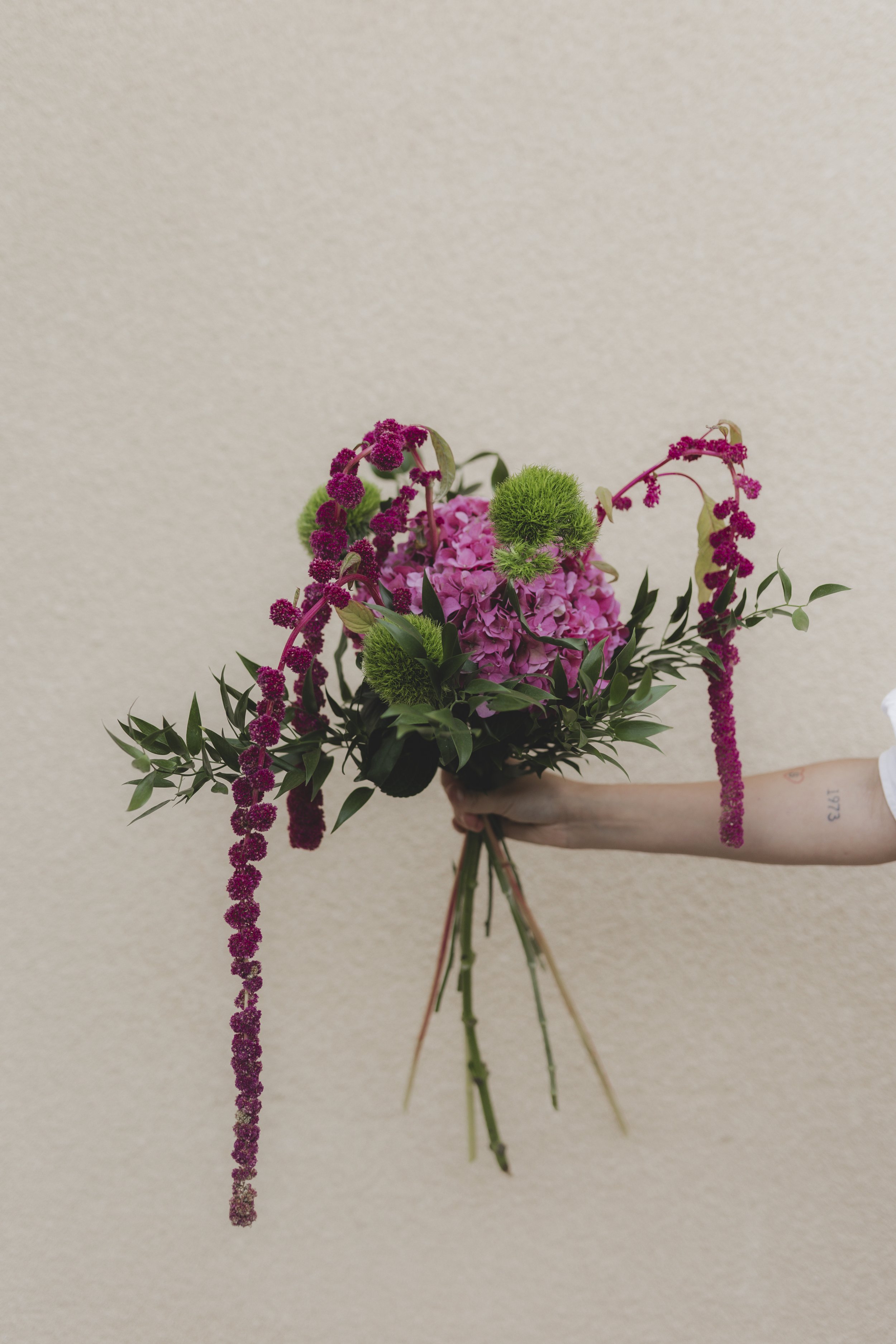 En hand håller ett blombud med rosa och gröna blommor mot en beige vägg.
